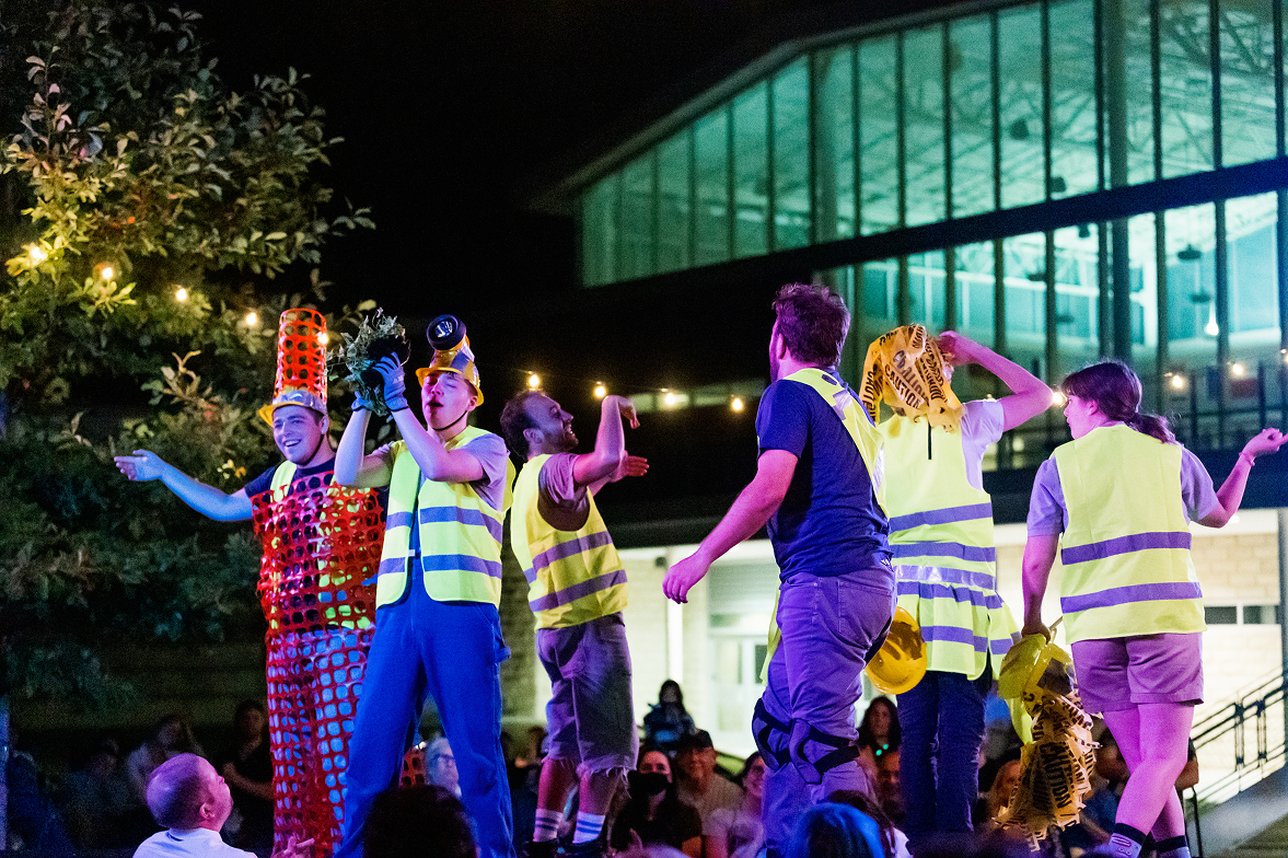 People dressed in safety vests and colorful costumes performing on an outdoor stage at night, with an audience watching.