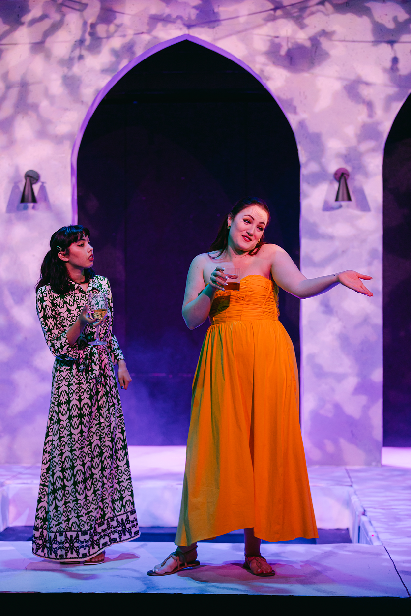 Two women stand on stage, holding drinks. One woman, in a yellow strapless dress, speaks or gestures, while the other, in a black and white patterned dress, listens.