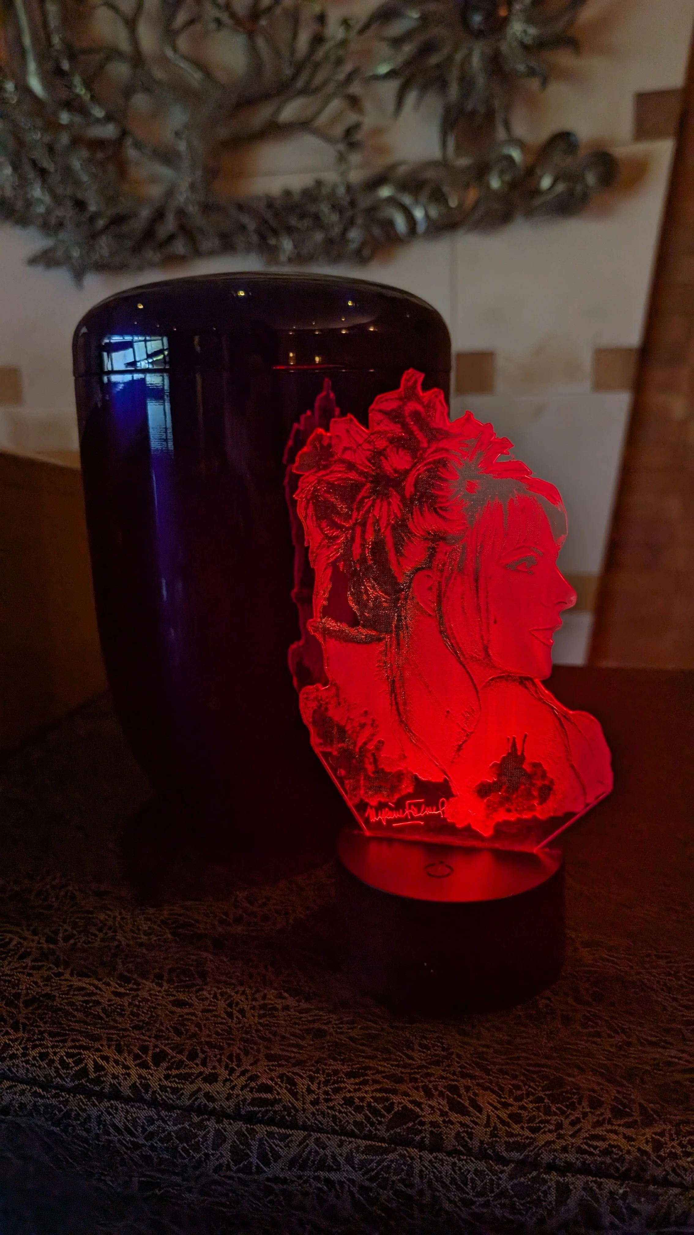 Une lampe LED rouge représentant un profil féminin avec des cheveux ornementés et des éléments de paysage. En arrière-plan, une urne noir cylindrique. La scène se déroule sur une nappe en dentelle noire.