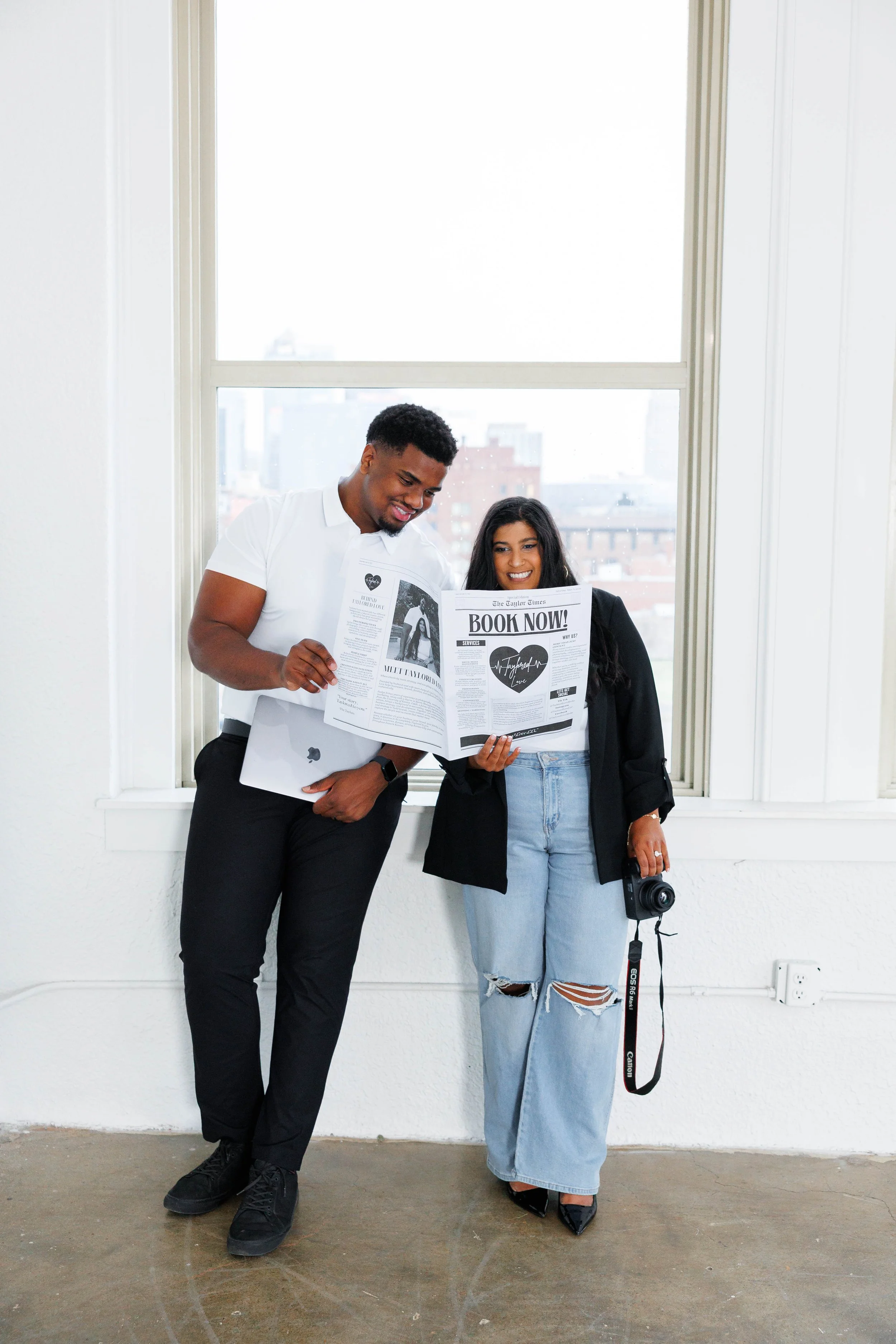 A young man and woman are standing by a large window, reading a newspaper together. The man is smiling and holding a laptop, while the woman is smiling and holding a camera. The woman is wearing ripped jeans and heels, and the man is dressed in black pants and shoes.