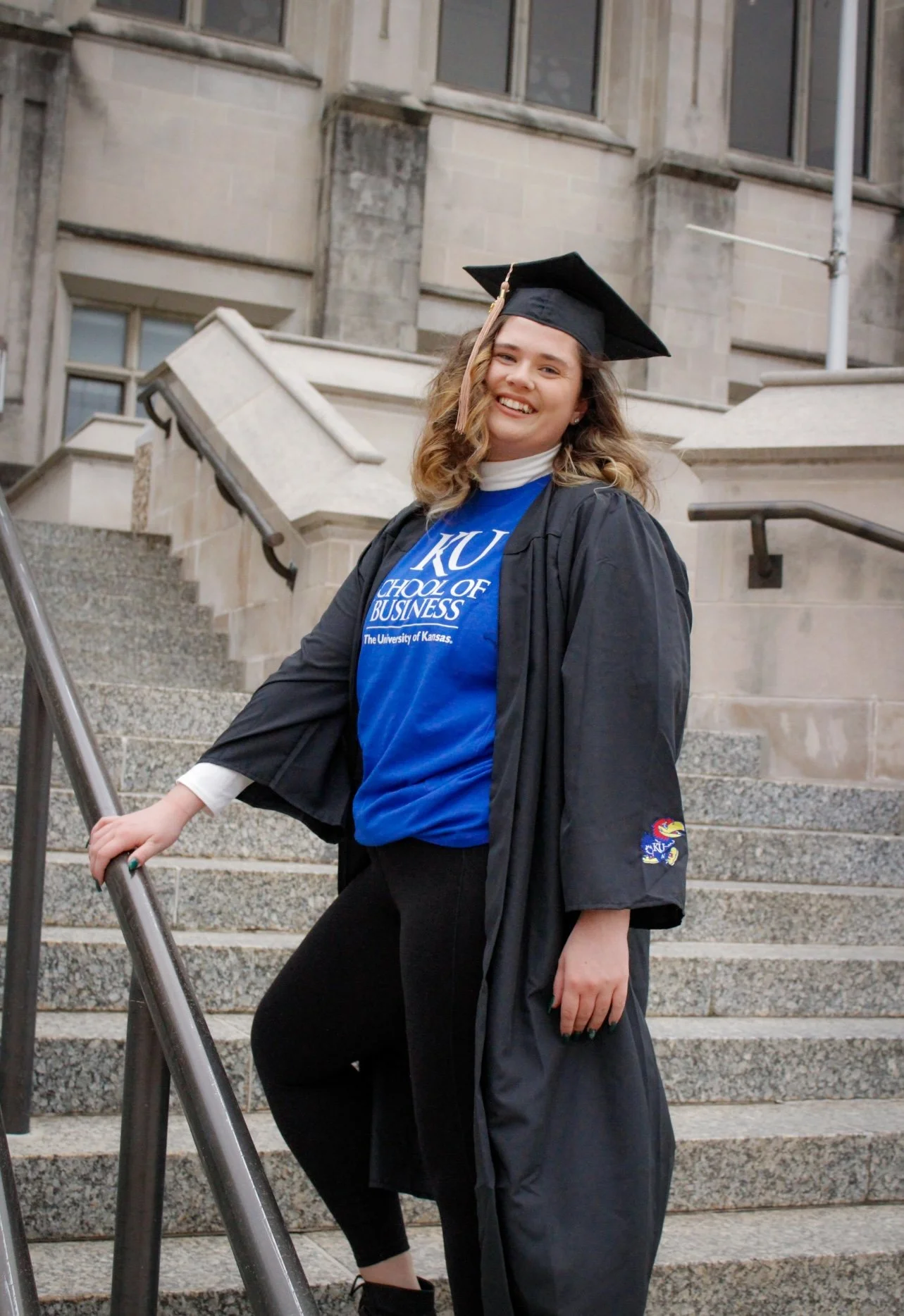 A young woman in a graduation cap and gown standing on outdoor steps, smiling, with a University of Kansas shirt visible.
