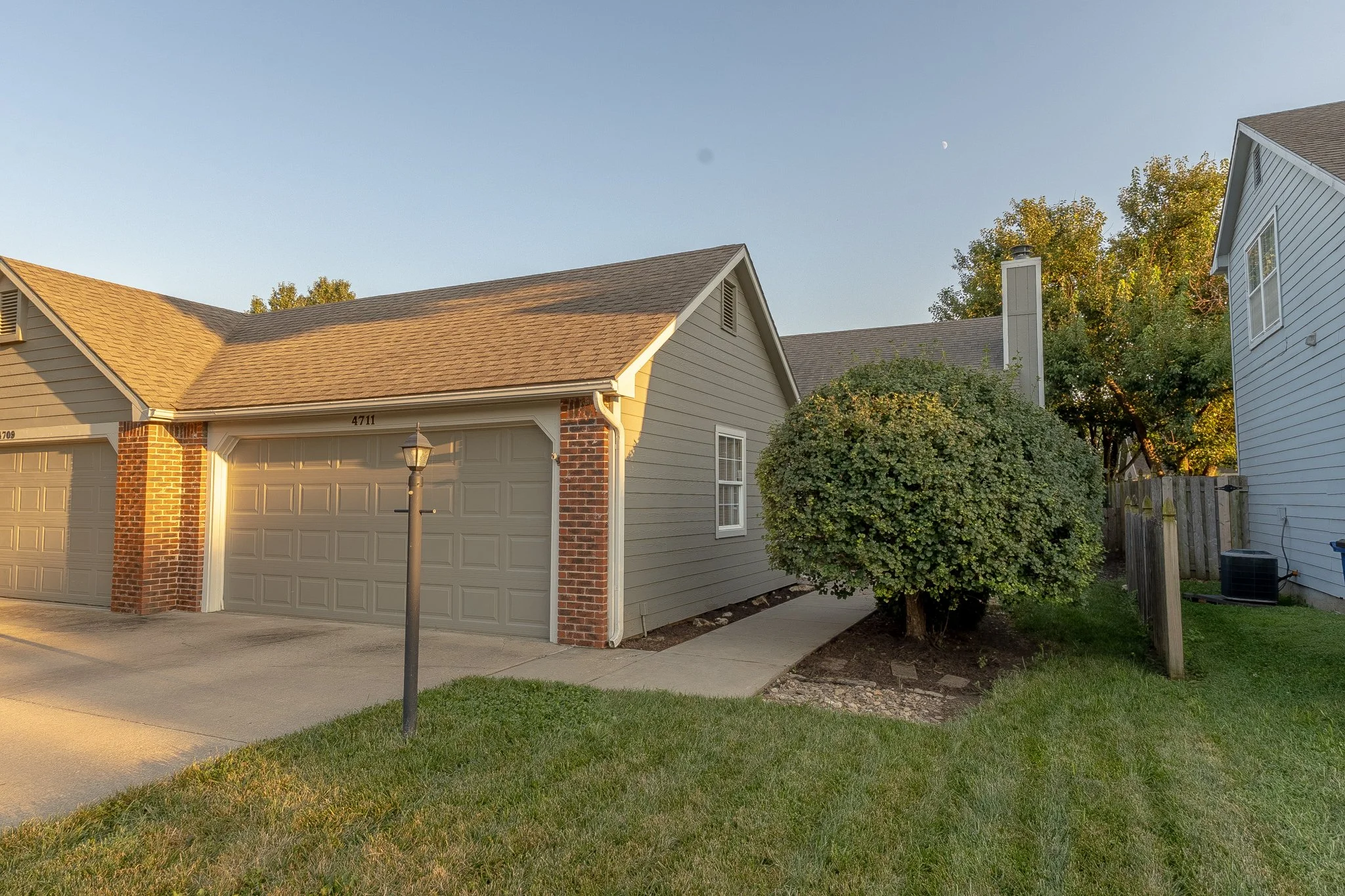 A suburban house with beige siding, a two-car garage, and a landscaped front yard with a small tree and grass. The sky is clear with a visible crescent moon.