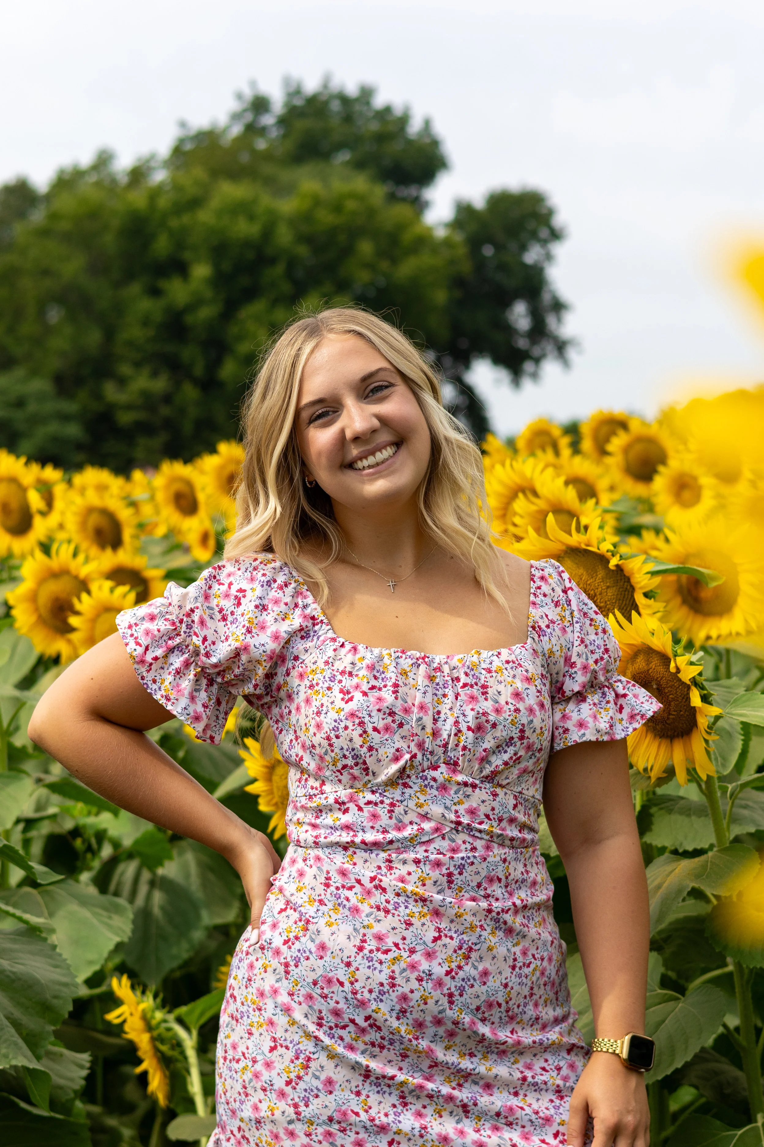 A smiling young woman with blonde hair wearing a floral dress standing in a sunflower field with trees and an overcast sky in the background.