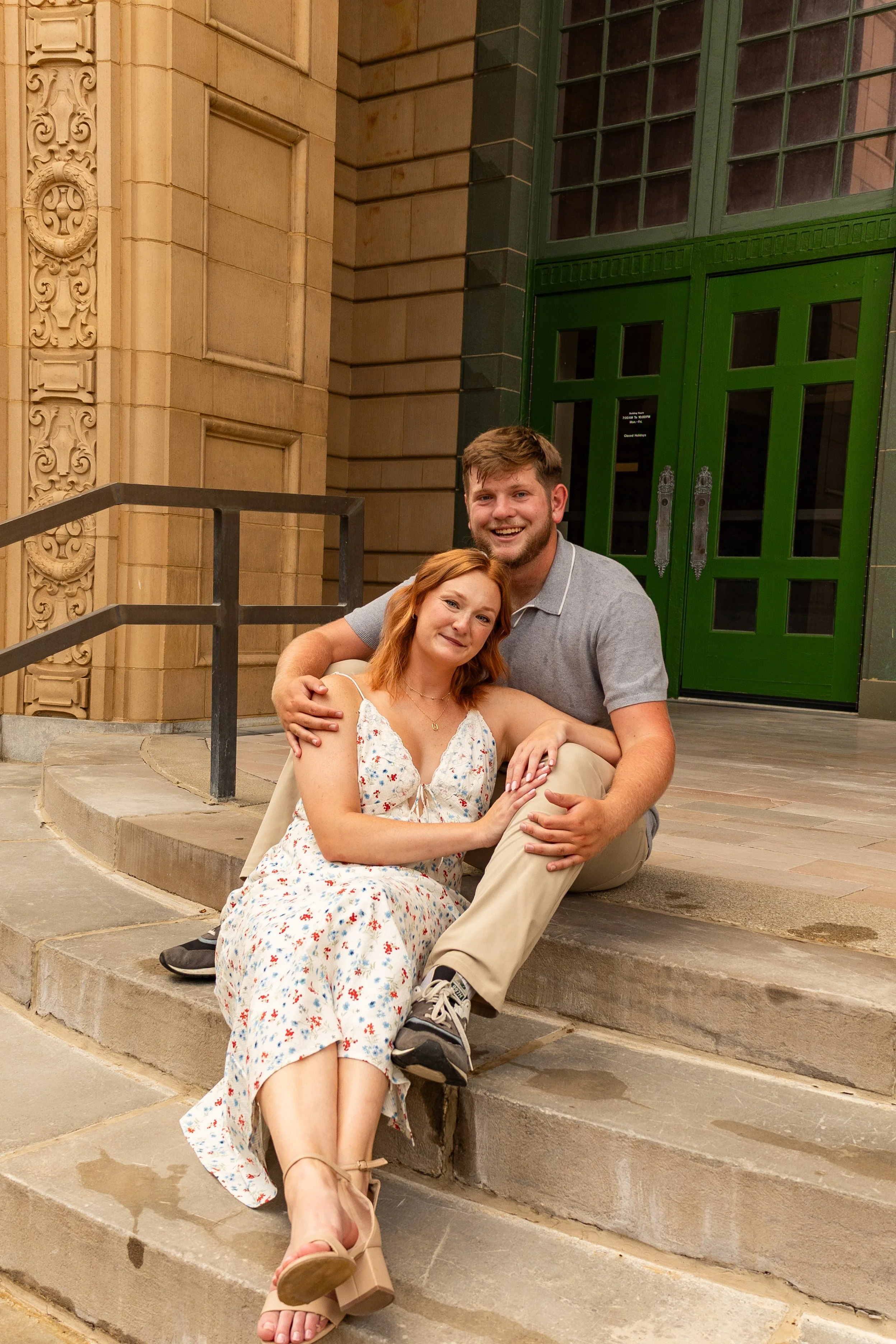 A young couple sitting on the steps outside a building with ornate architectural details. The woman has red hair and is wearing a white floral dress and beige sandals. The man has brown hair and is wearing a gray polo shirt, khaki pants, and gray sne