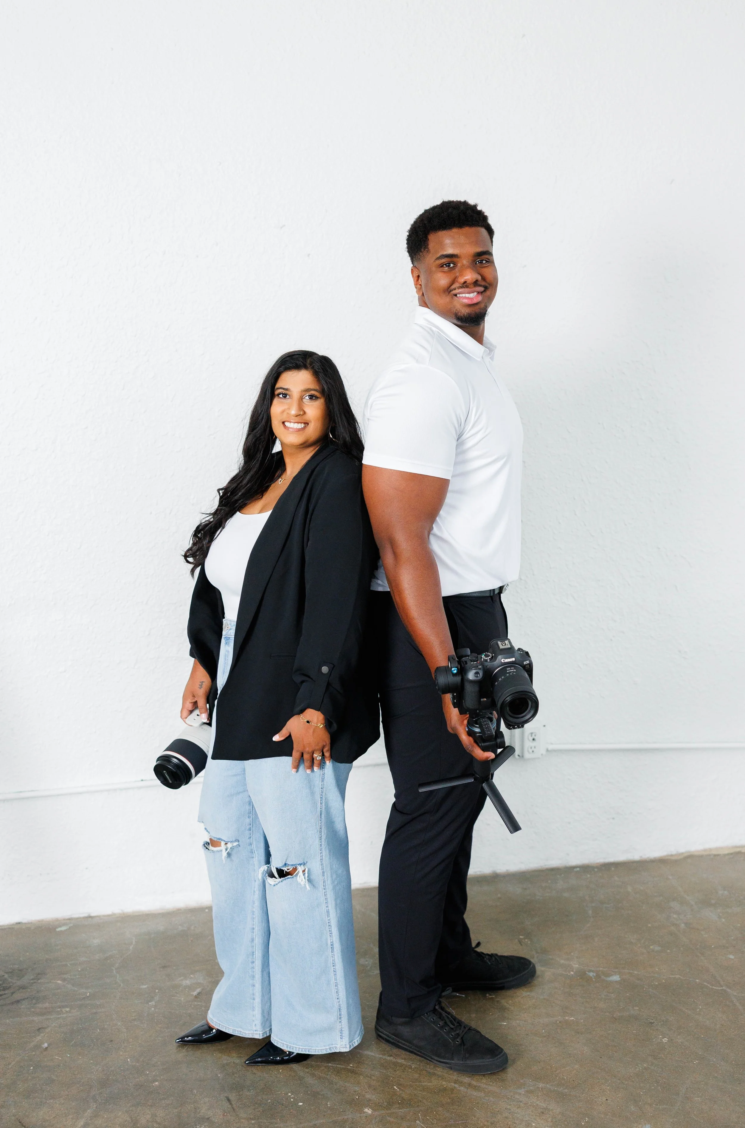 Professional branding photography picture showing a man and a woman standing against a plain white wall, both holding professional camera equipment, with the man holding a DSLR camera on a tripod and the woman holding a telephoto lens, both smiling.