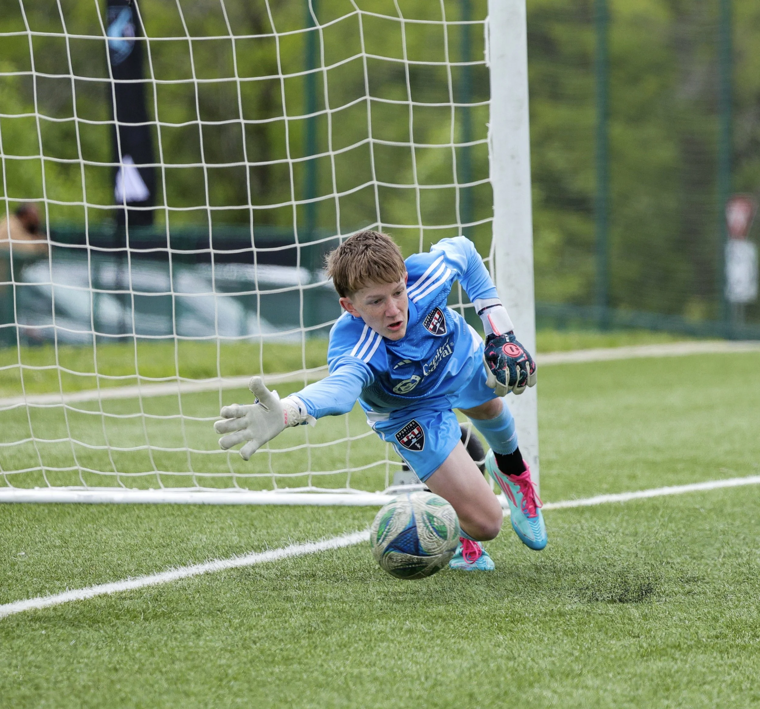 A young soccer player in a blue jersey and pink shoes dives to save the ball in front of the goal, with a goalkeeper glove visible and a goalpost behind him, on a soccer field.
