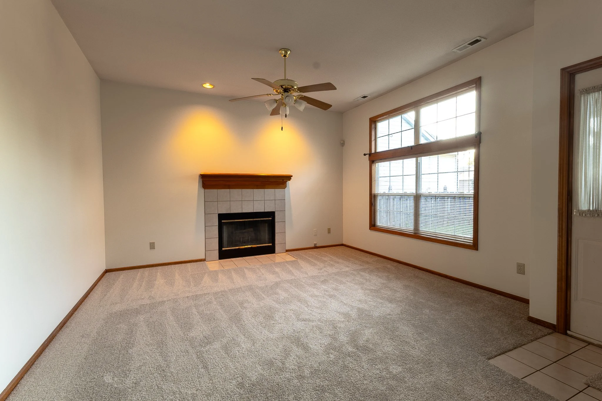 Empty living room with a fireplace, carpeted floor, large window with blinds, and ceiling fan.