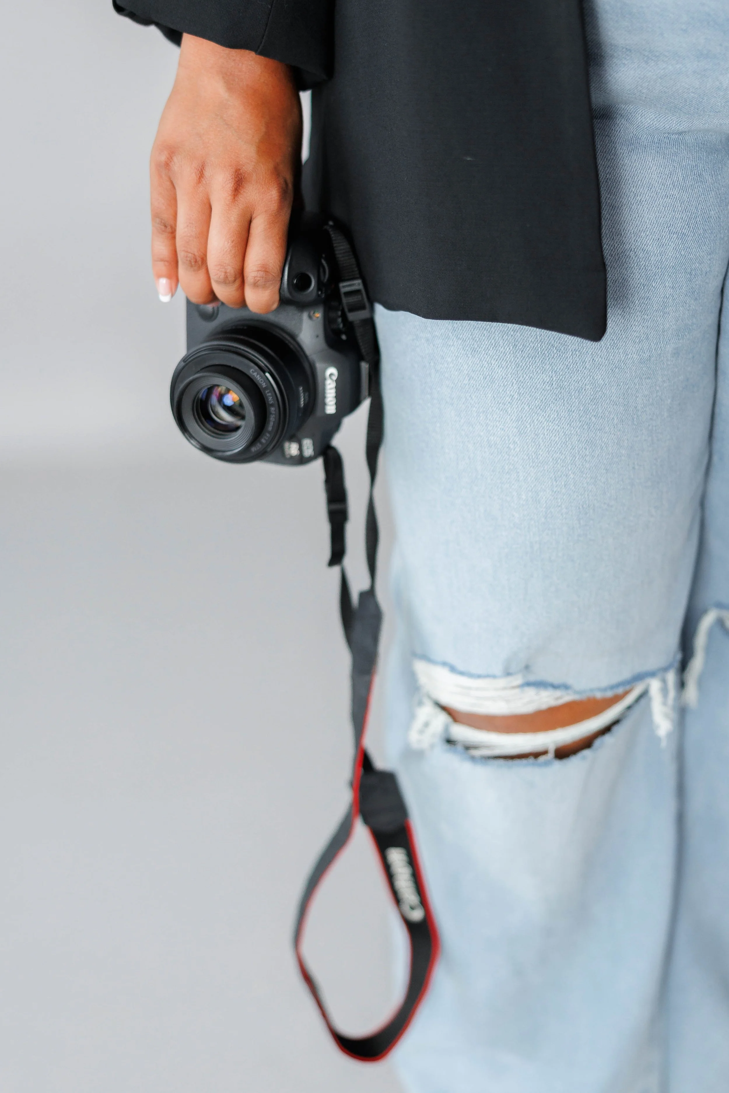 Person holding a Canon camera with a strap, wearing a black and gray outfit and ripped denim shorts.