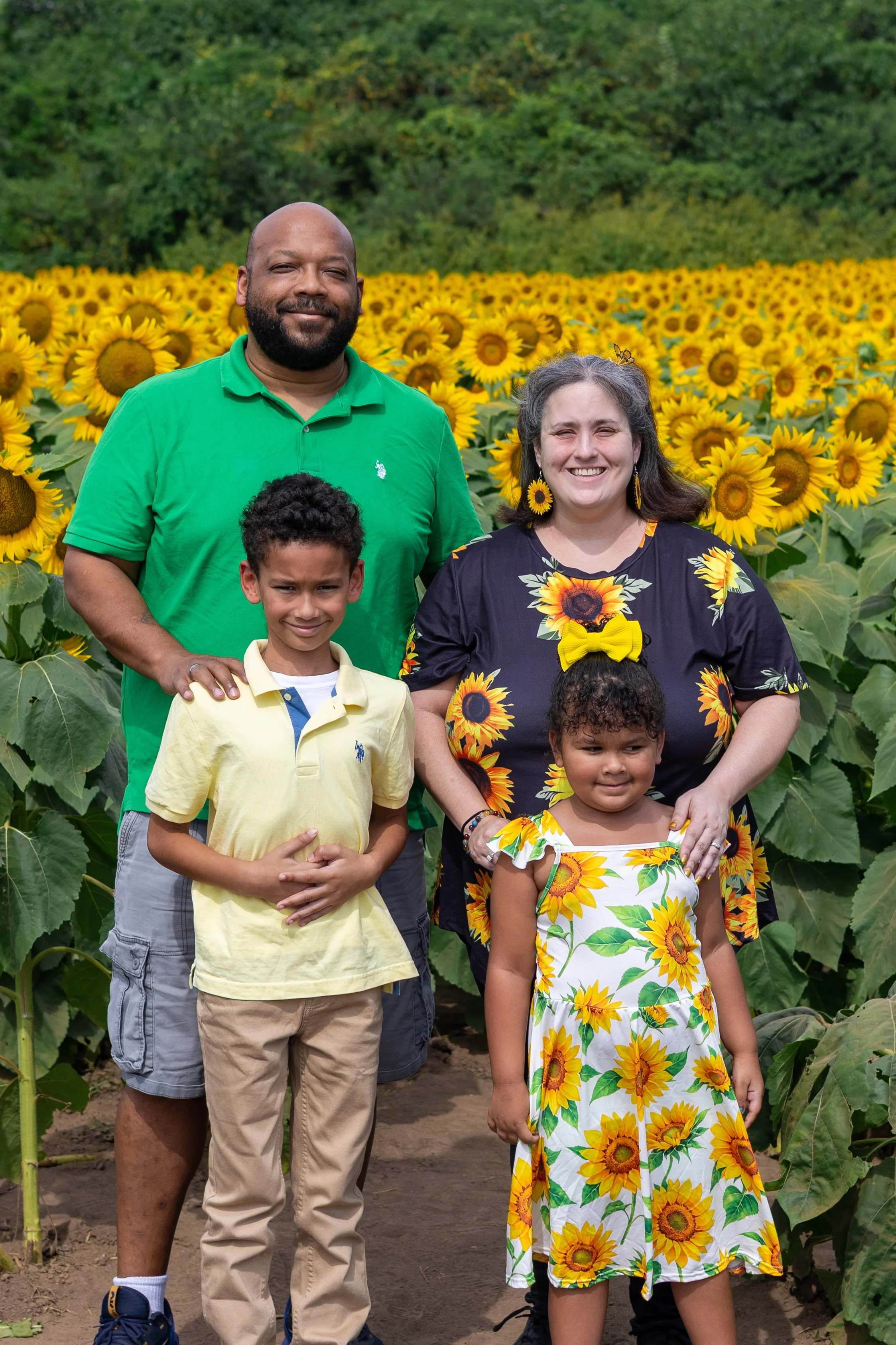 A group of six people standing in a sunflower field with a green, tree-covered hillside in the background.