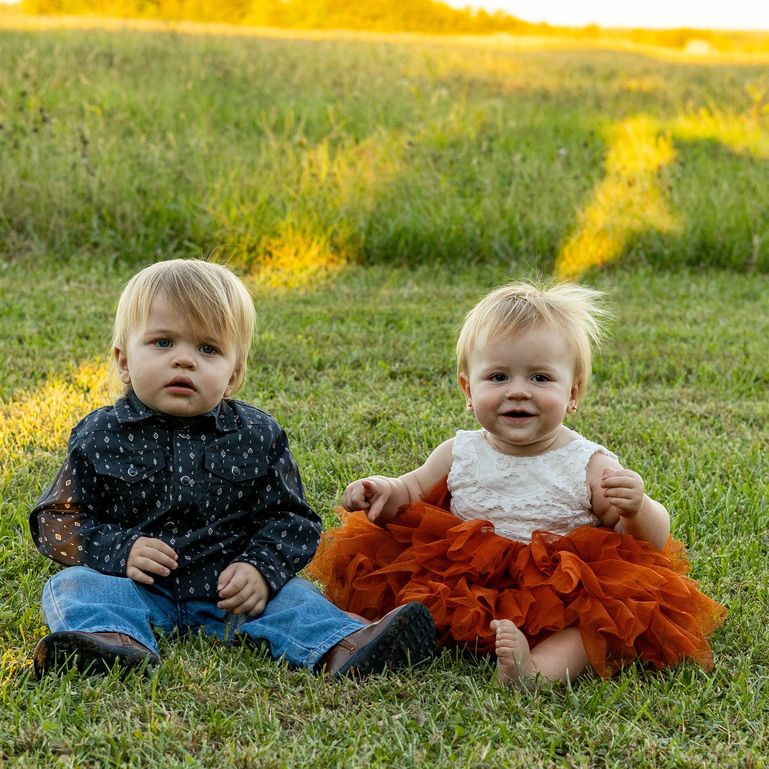 Three young children sitting on grass outdoors during sunset, two girls and a boy.
