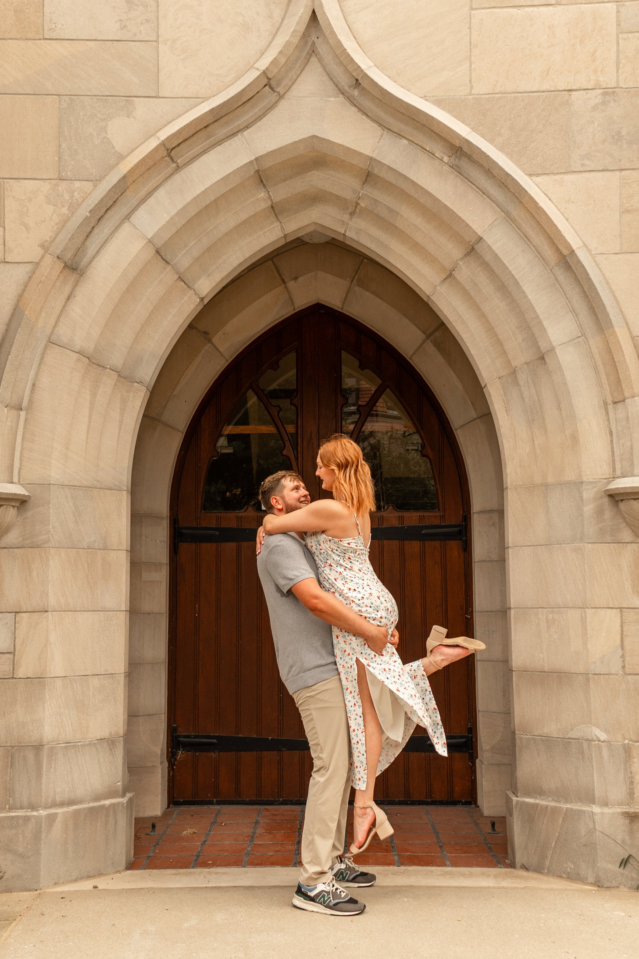 A smiling man lifting a woman in a floral dress in front of a stone arch gateway, with a wooden door behind them and brick pavement underneath.