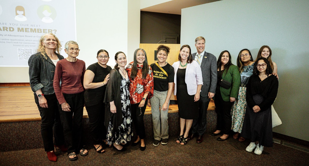 Group of 12 diverse people standing on a stage in a conference room, smiling for a photo.