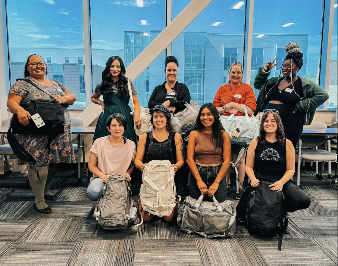 Group of nine diverse women, some with backpacks, posing in an indoor space with large windows and city buildings in the background.