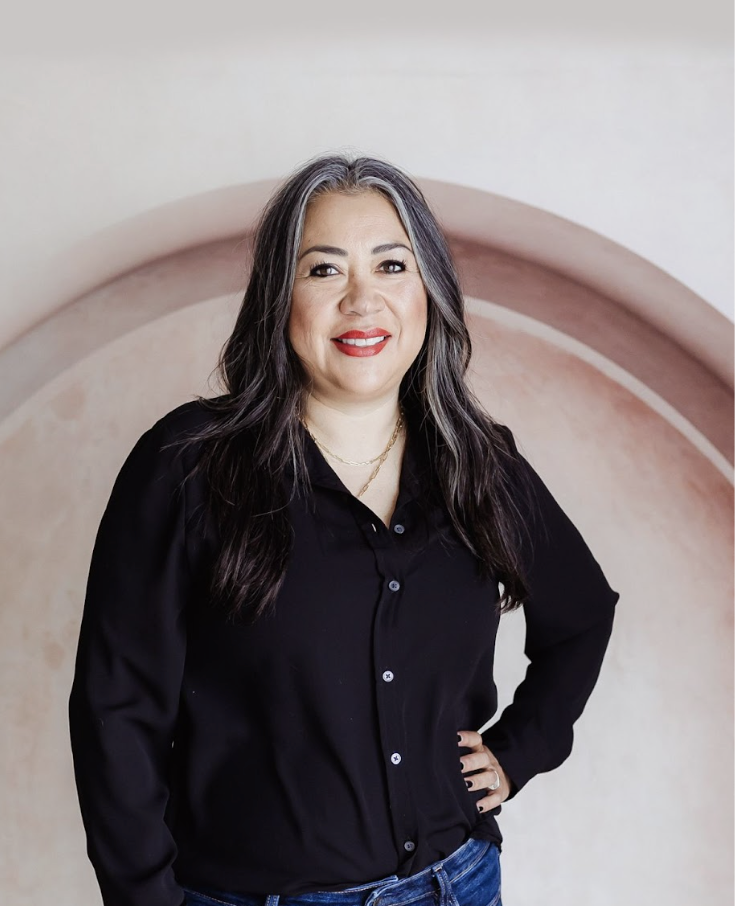 A woman with long dark hair and gray streaks, smiling and wearing a black button-up shirt and blue jeans, standing in front of a circular pink and beige background.