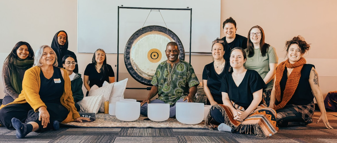 Group of people sitting and kneeling in a circle indoors, with a gong and singing bowls in front of them, smiling at the camera.