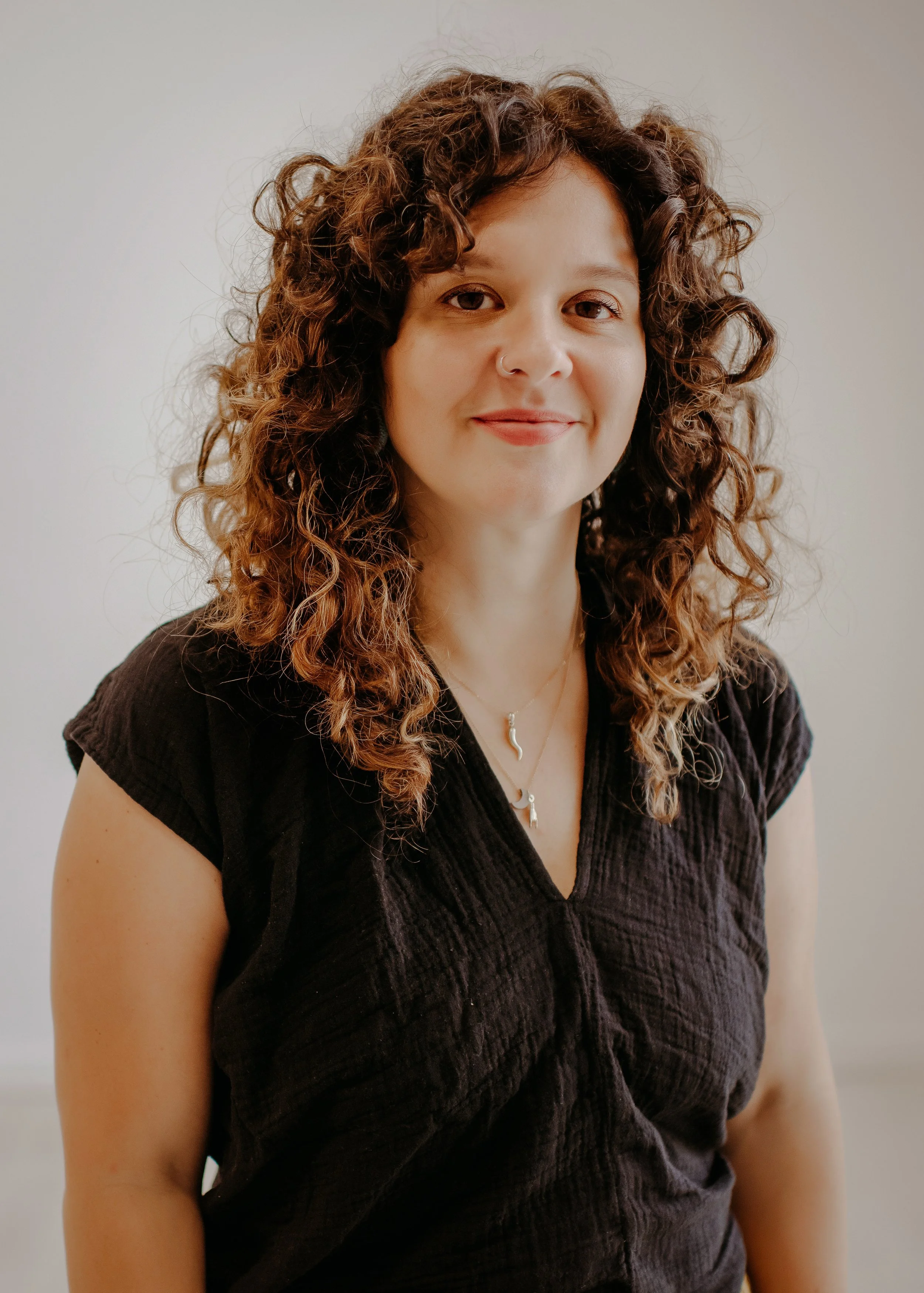 A woman with curly brown hair, wearing a black top and layered necklaces, smiling at the camera.