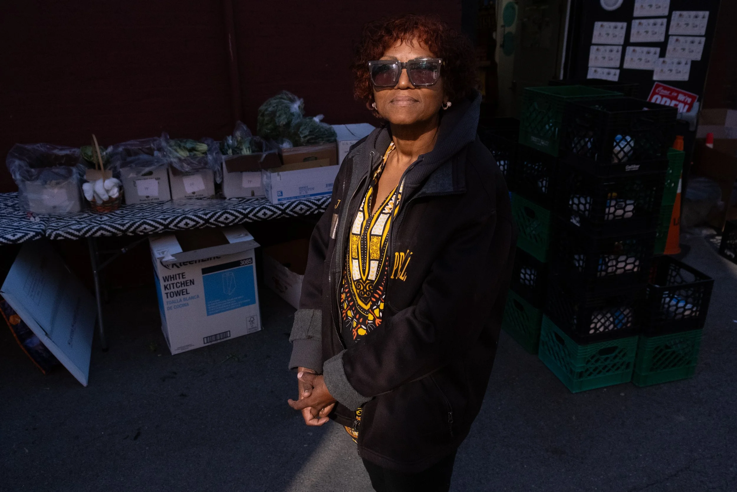 Jackie LaSonde stands outside of the food pantry wearing large sunglasses and a black jacket with a sliver of sunlight peaking on her face.