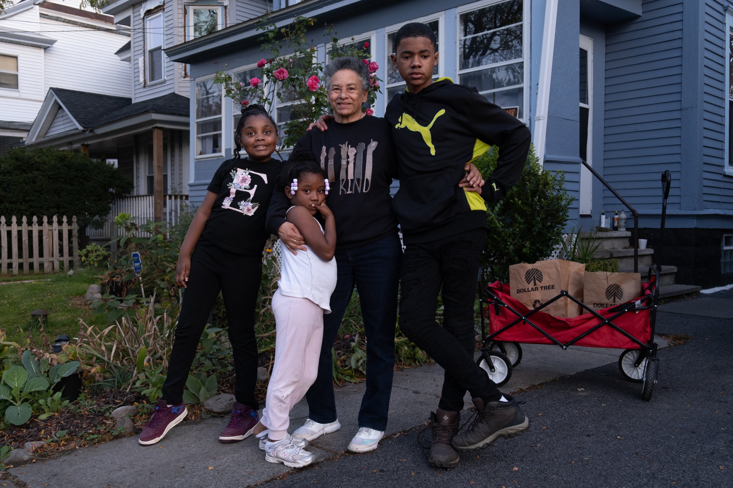 Rhonda and her three grandchildren pose for a photo outside of her blue home and a rose bush nearby.