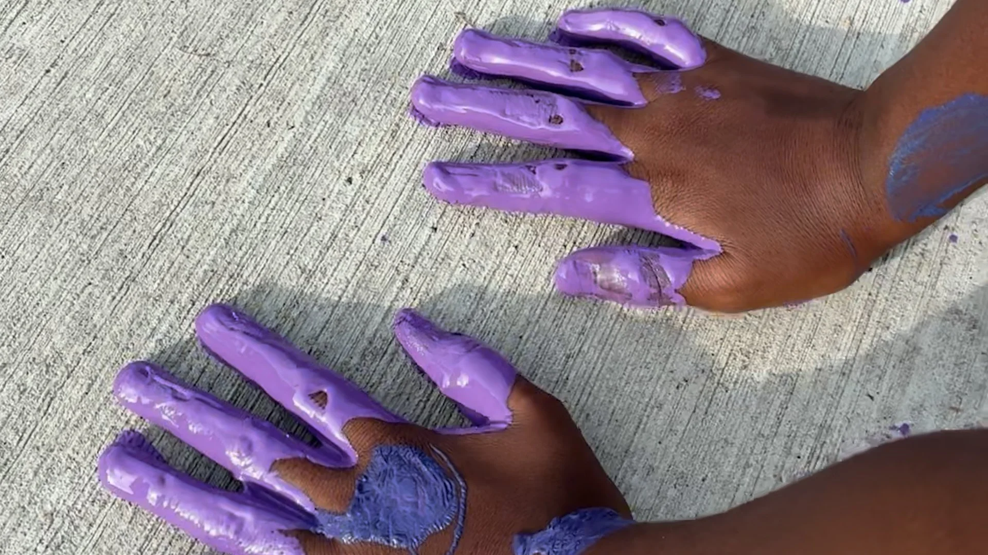 Two hands with purple paint on them stamping a hand print on the sidewalk.