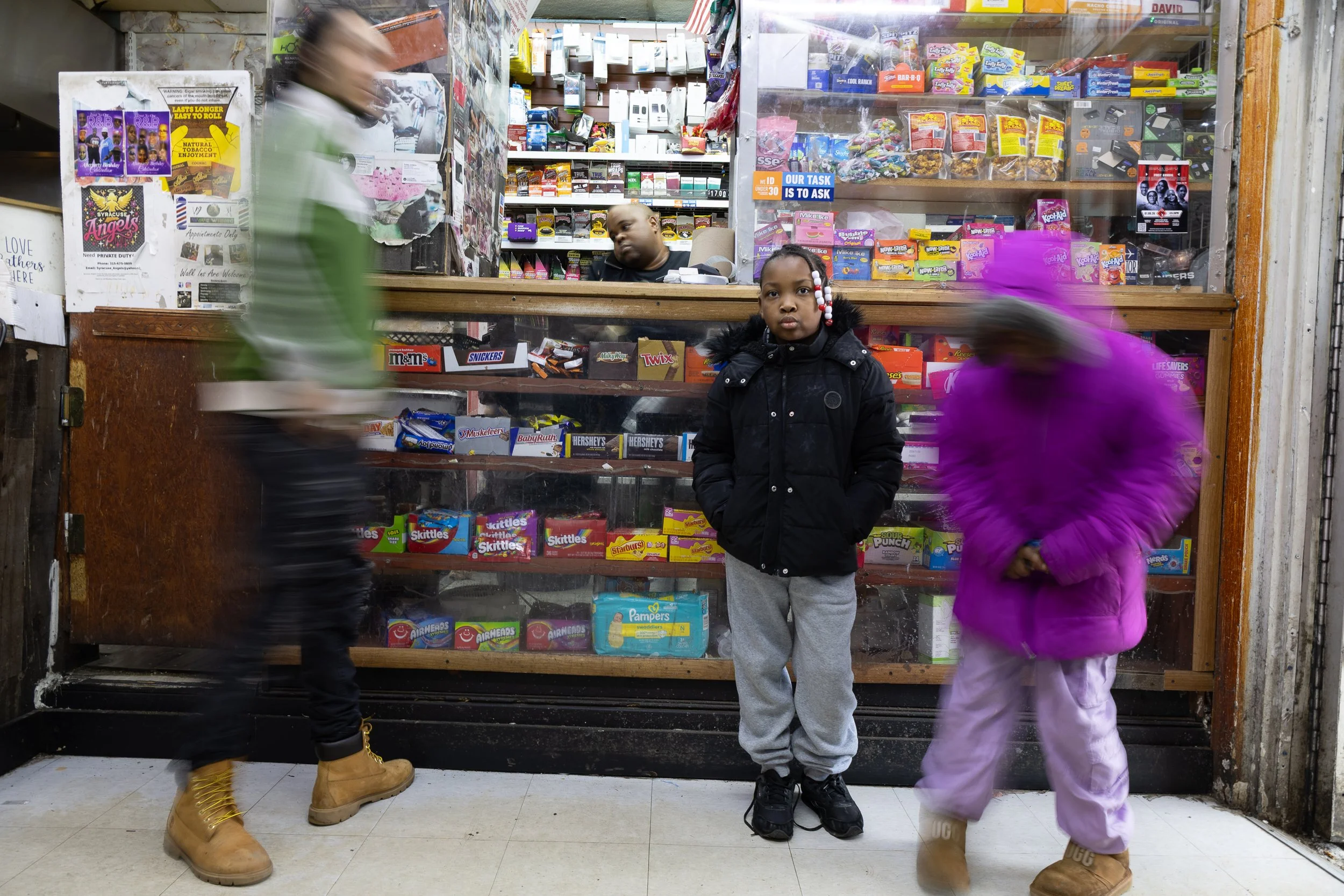  Two children pose in front of the counter at Ze Mart on Jan. 10, 2026, one blurred by motion; an adult approaches the counter at left and Kelvin Jackson is visible in the background. Photo by: Destinyi Fernandez 