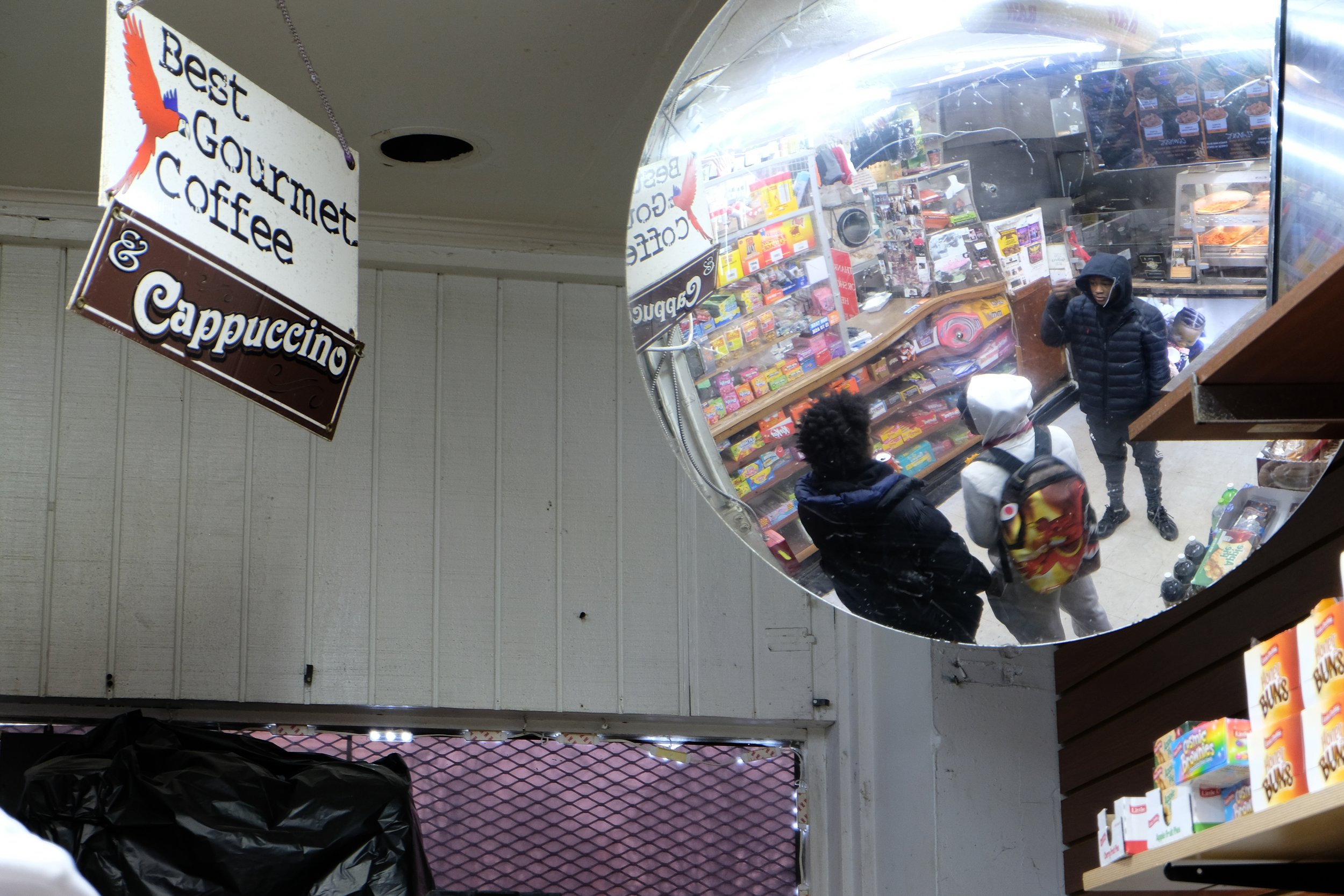  Teens are reflected in a convex corner mirror inside Ze Mart while waiting at the counter, with a sign advertising coffee and cappuccino visible at left, on Jan. 10, 2026. Photo by: Micah Orieta 