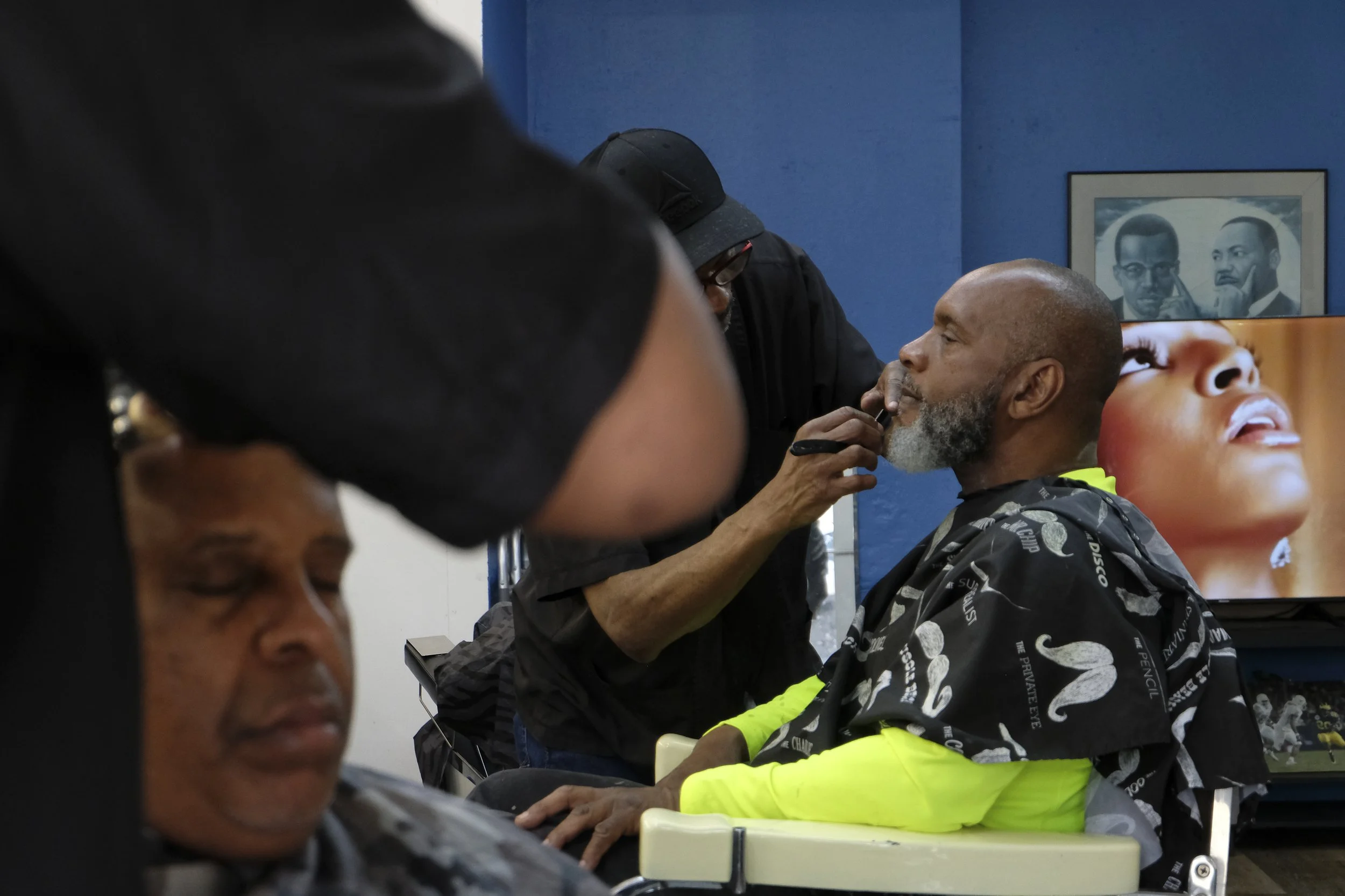  Derrick Franklin's arm frames a patron receiving a head shave in the foreground, while barber David Blue works on another patron's beard in the background, at Blue Brothers Barber Shop on South Salina Street in Syracuse, May 16, 2026. Photo by Zahra
