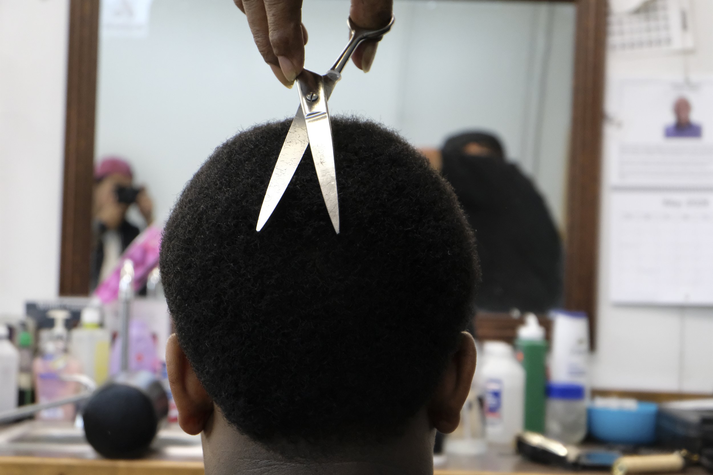  Scissors and Derrick Franklin's hand enter the frame above a patron's head, seen from behind, at Blue Brothers Barber Shop on South Salina Street in Syracuse, May 16, 2026. Photo by Zahra Perkins. 
