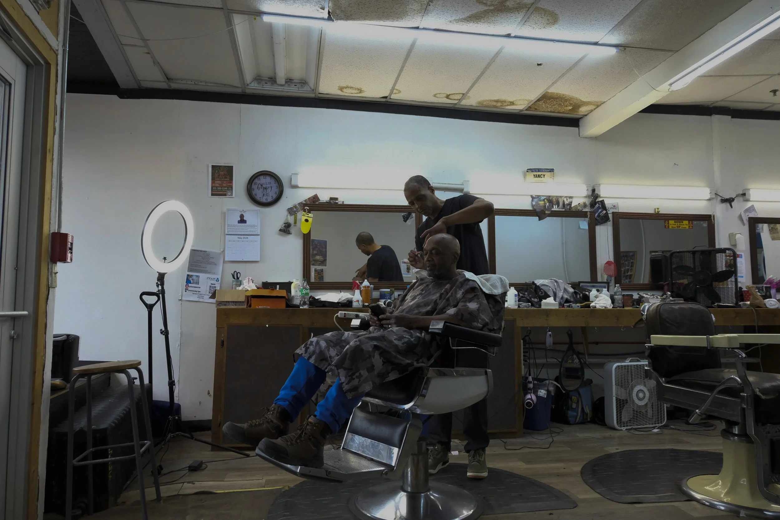  A wide shot shows Dexter Bullock working on a patron in the chair at Blue Brothers Barber Shop on South Salina Street in Syracuse, May 23, 2026. Photo by Zahra Perkins. 