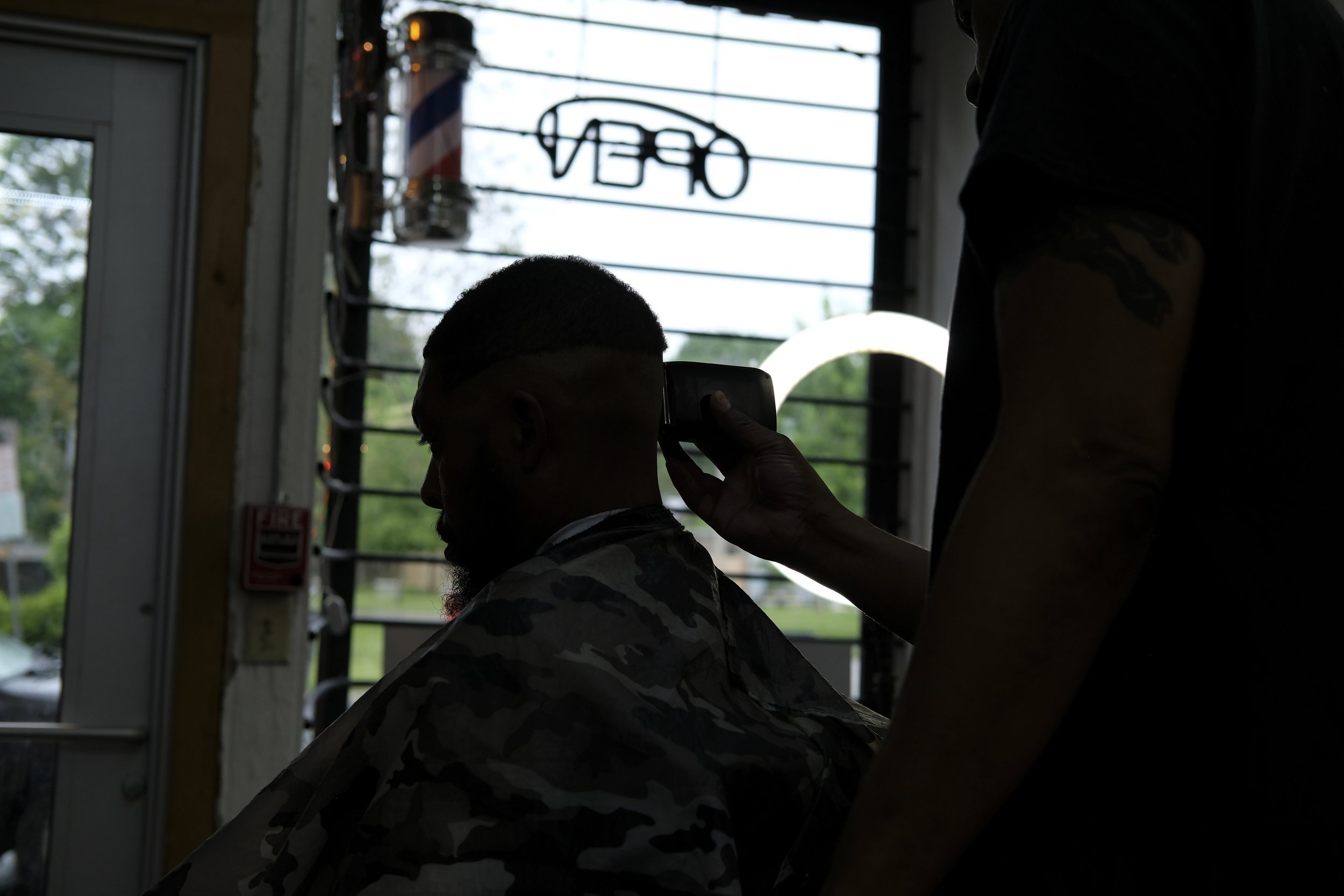  Dexter Bullock and a patron appear in silhouette against a window overlooking South Salina Street, with the shop's barber pole and "Open" sign visible, at Blue Brothers Barber Shop in Syracuse, May 23, 2026. Photo by Manila Southammavong. 