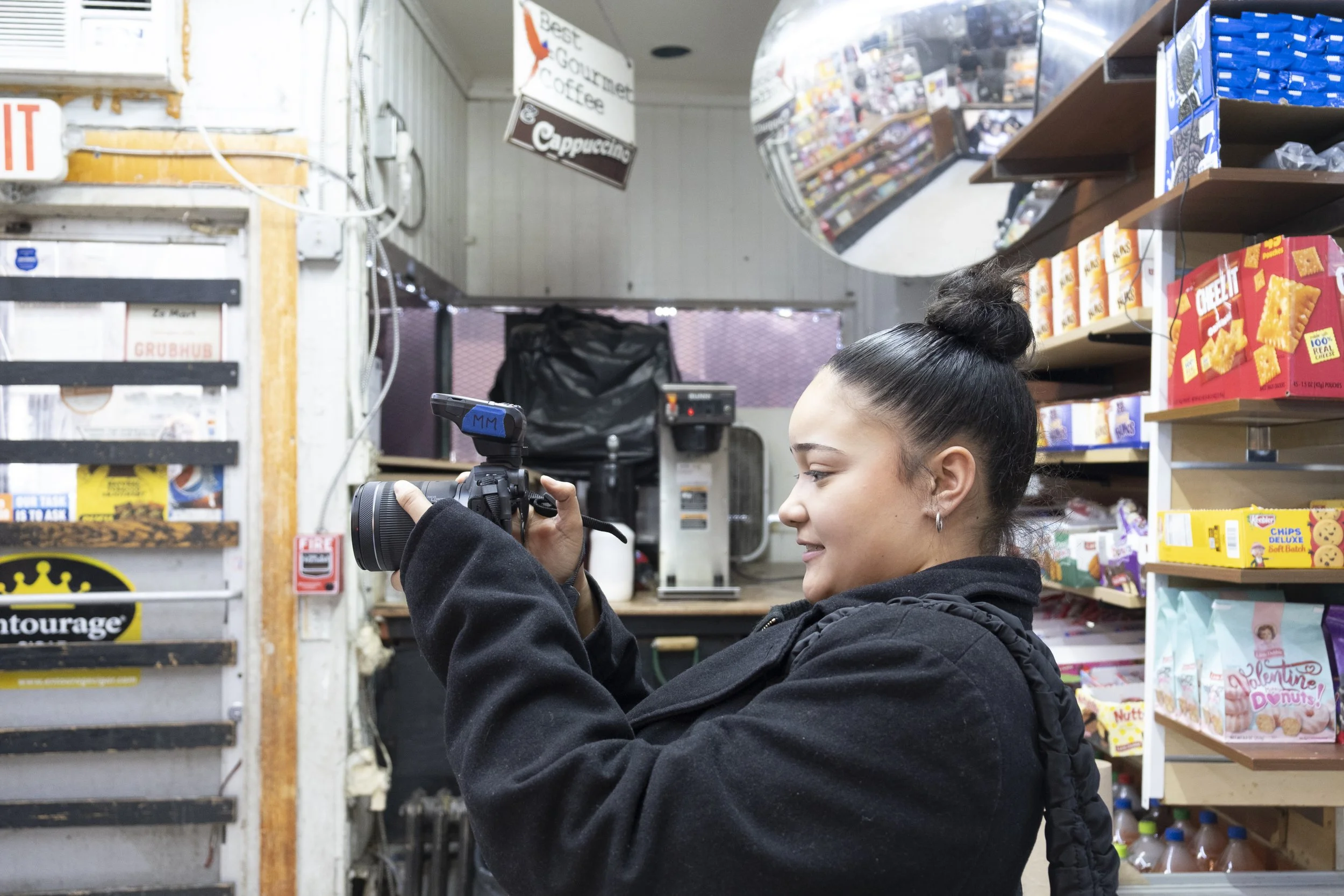 A young woman with dark hair tied in a bun wearing a black hoodie, standing in a convenience store, holding a camera, and smiling while taking a photo.