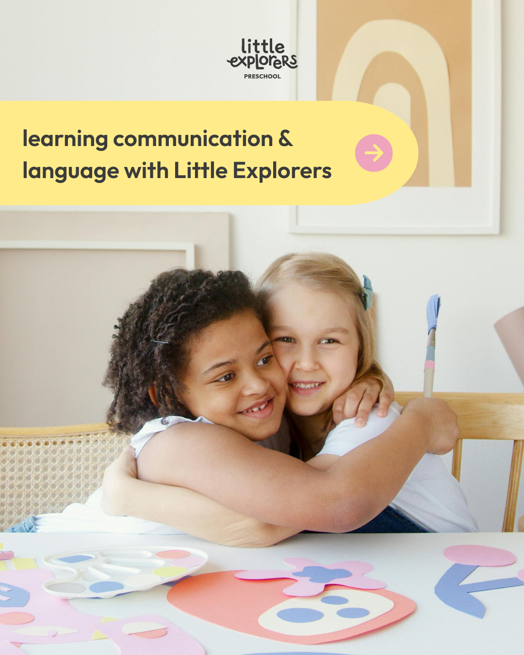 Two young girls hugging each other at a table decorated with paper craft art projects, with a colorful abstract art piece on the wall behind them.