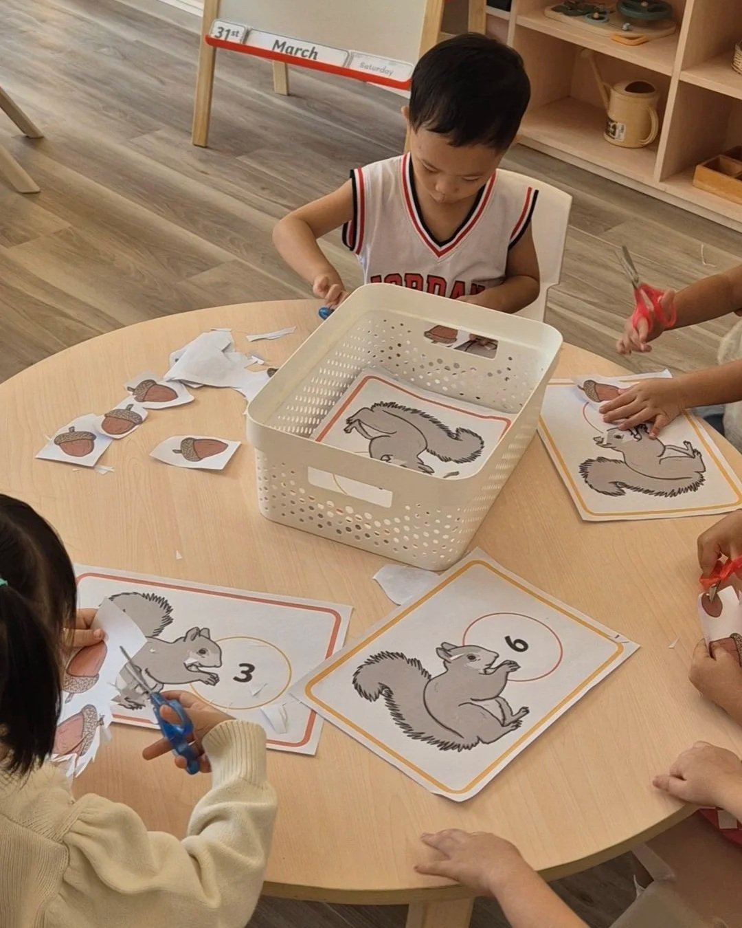 Children cutting and matching pictures of squirrels with numbers on a classroom table.