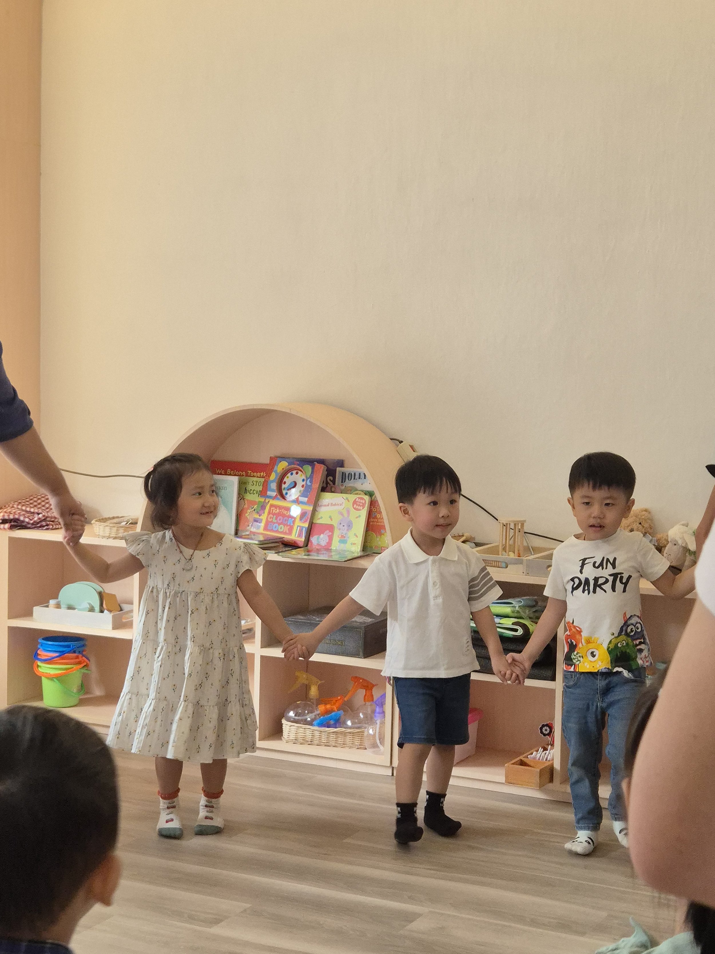 Three young children are holding hands in a circle, standing in a classroom or playroom with shelves filled with books and toys in the background.