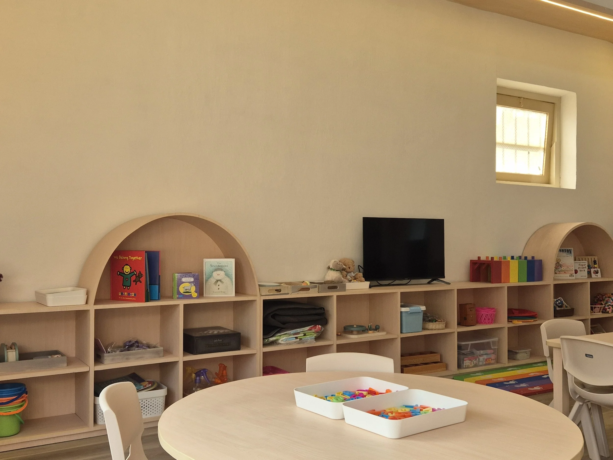 A preschool classroom with round light-colored tables and white chairs, with educational toys and books on wooden shelves, including plush toys, puzzles, and art supplies. There is also a small TV on the top shelf and a window allowing natural light.