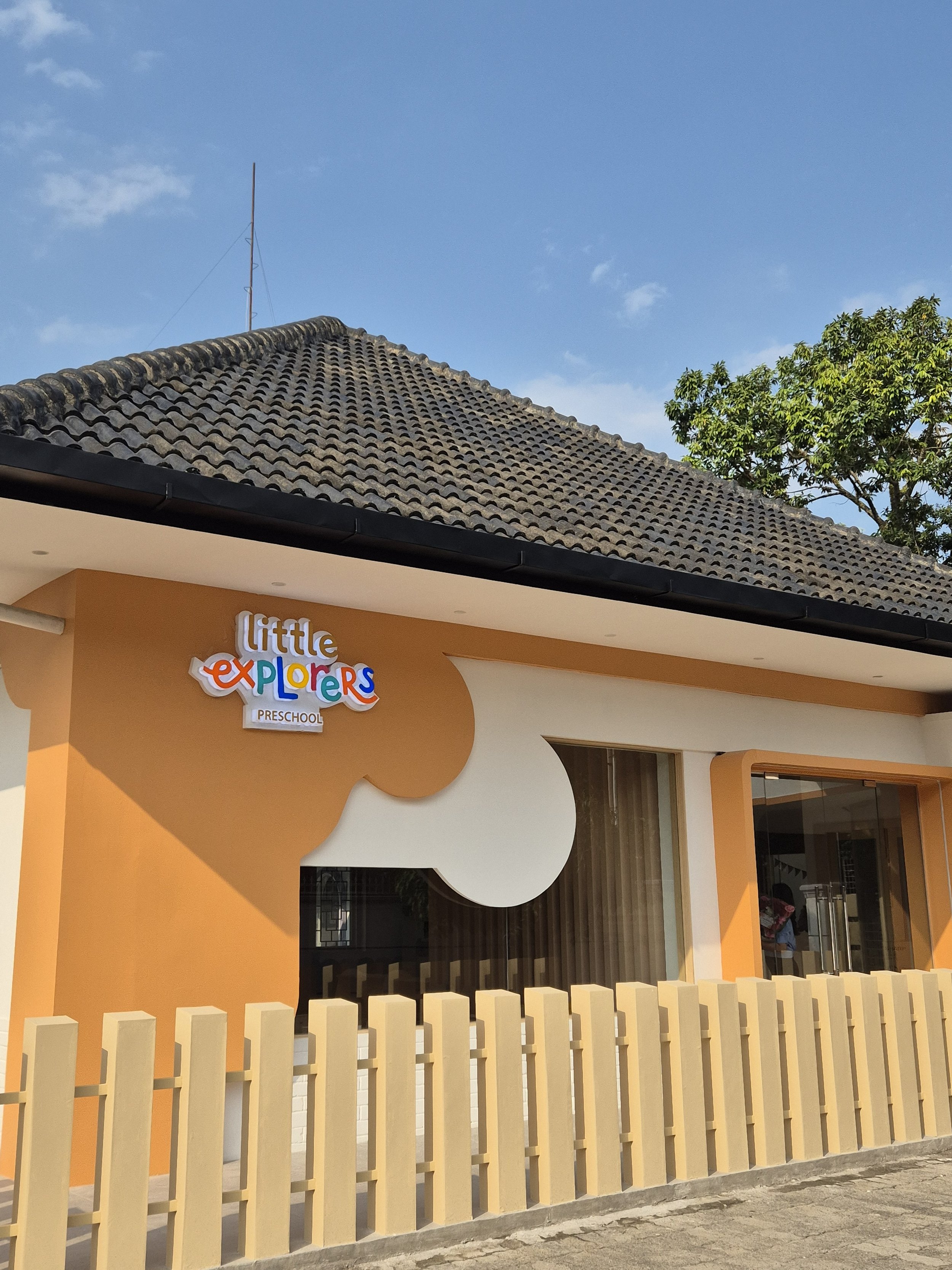 Exterior of a preschool called 'Little Explorers' with a colorful sign, orange and white walls, a wooden fence, and a brown tiled roof, under a blue sky with some clouds and a tree nearby.
