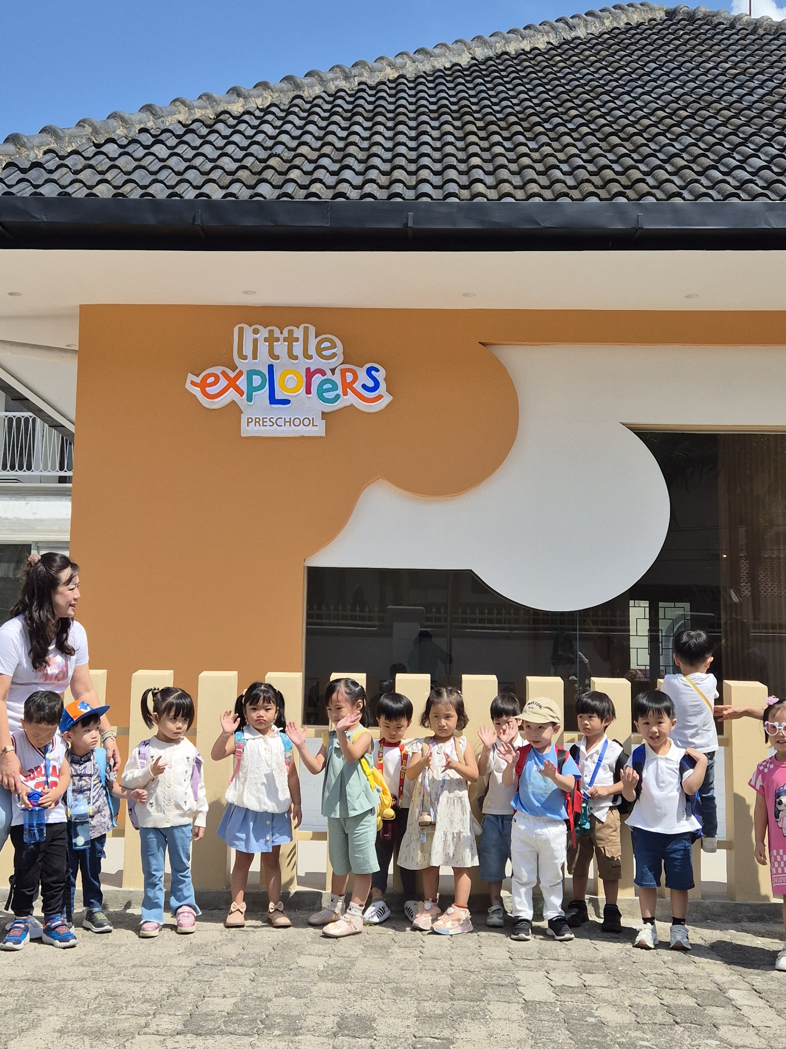 Group of young children standing outside a preschool named Little Explorers Preschool, with a teacher nearby, under a cloudy sky.