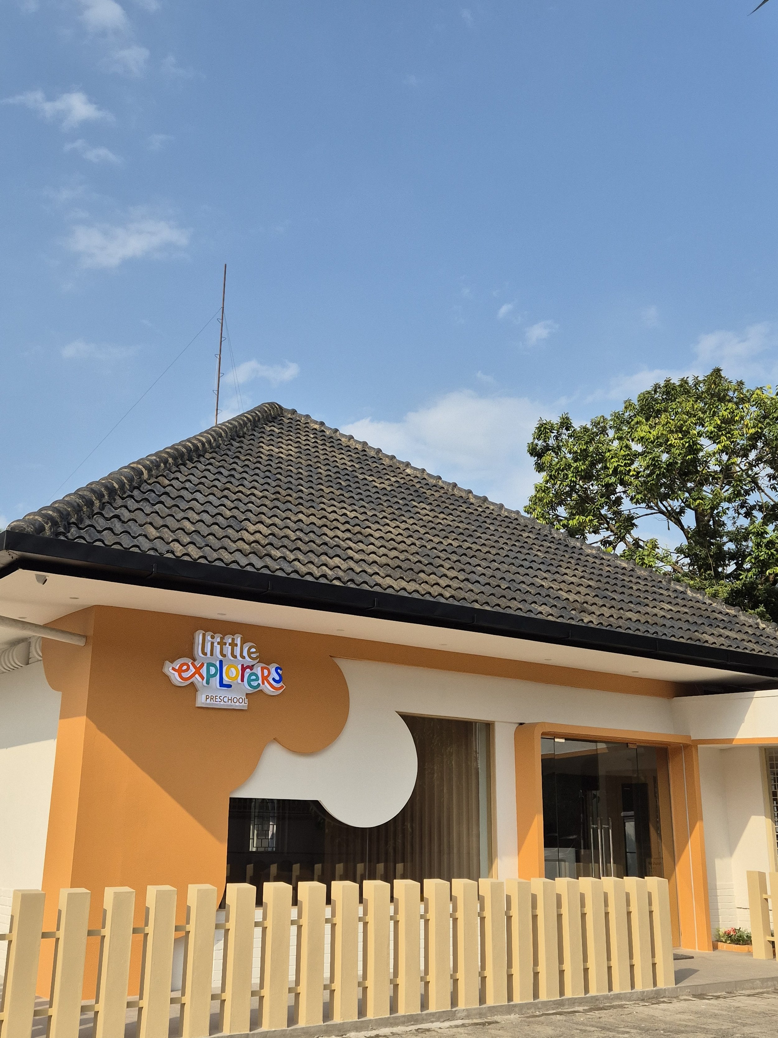 Exterior of a preschool building with a sign that reads 'Little Explorers Preschool', featuring a colorful, playful design, surrounded by a beige fence, under a clear blue sky with some clouds and trees.