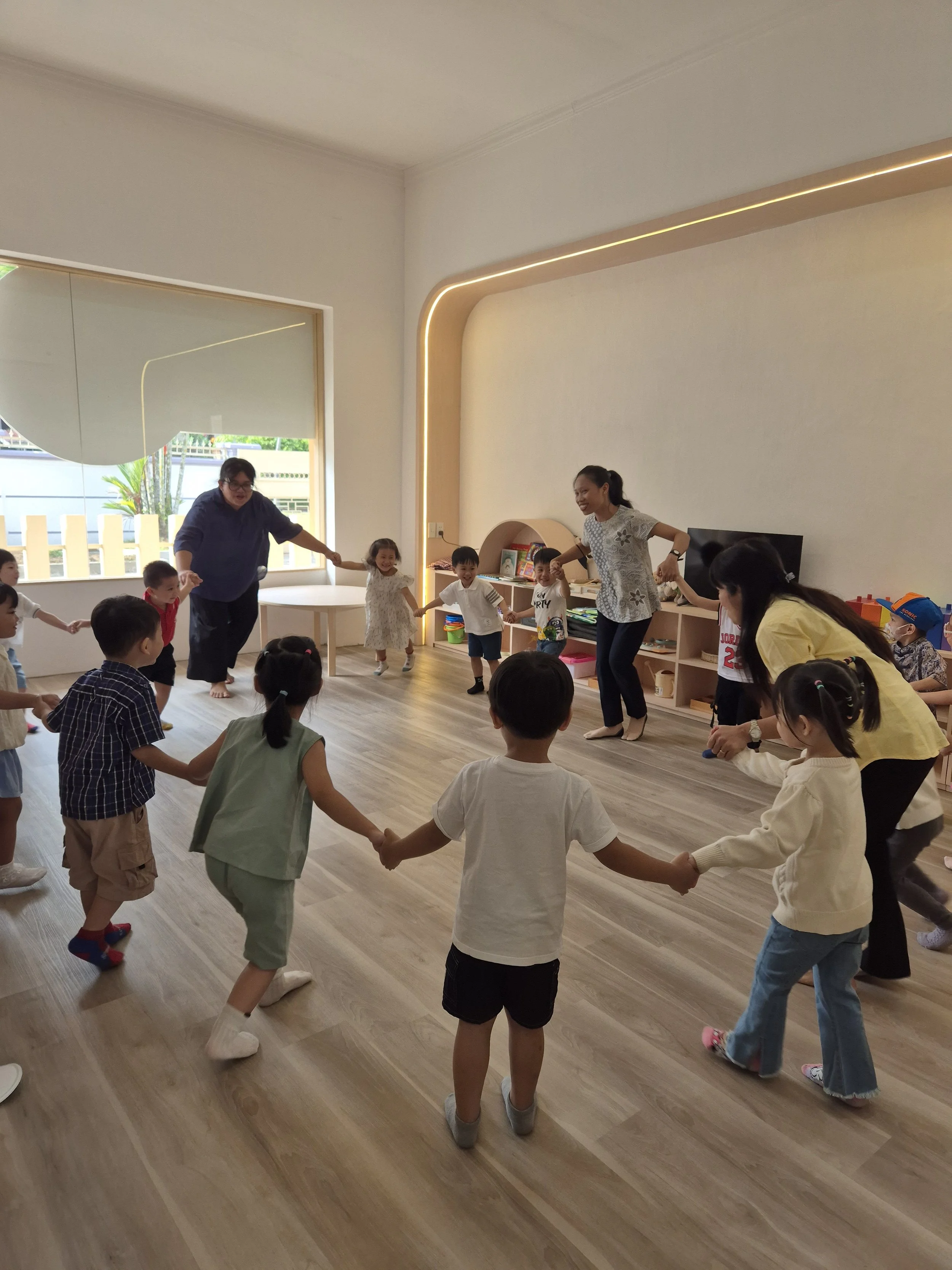 Children and teachers holding hands in a circle, playing inside a classroom.