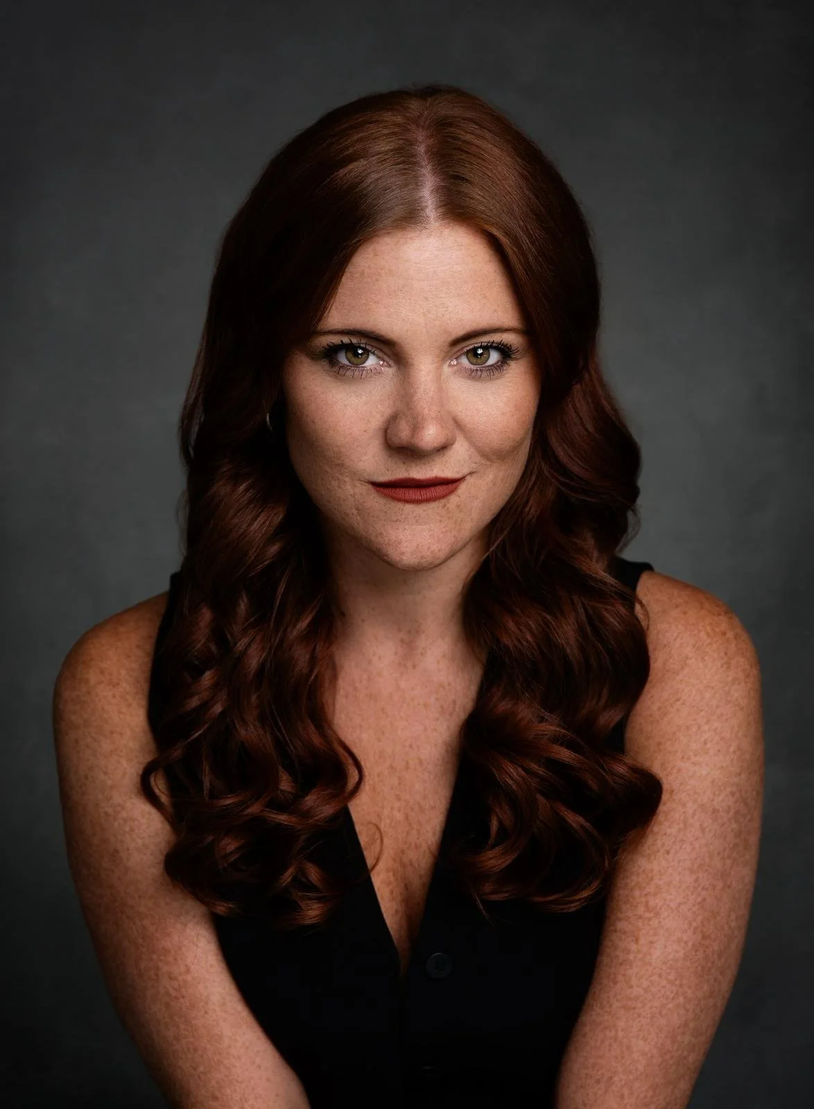 A young woman with long, wavy auburn hair and light skin with freckles, wearing a sleeveless black top, looking directly at the camera with a confident expression against a dark gray background.