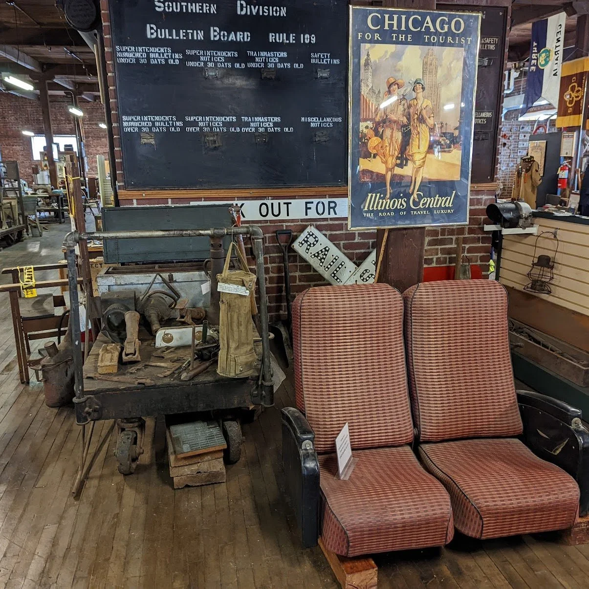 Vintage furniture and memorabilia including a vintage blackboard, a colorful poster promoting Chicago for the tourist by Illinois Central, an old train sign, pipe tools, and two red striped seats inside a rustic, brick-walled space with wooden floors