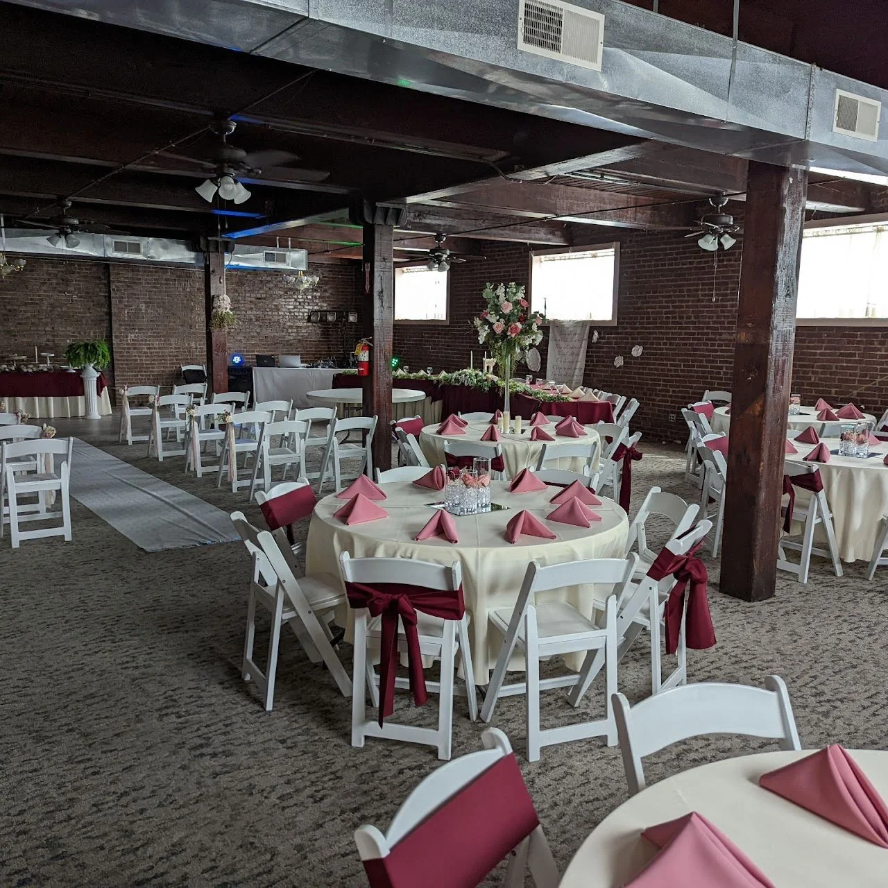 Event space decorated with round tables covered in cream tablecloths and white chairs with pink sashes, pink napkins on the tables, and a large floral centerpiece. There are brick walls, windows, and ceiling fans in the room.