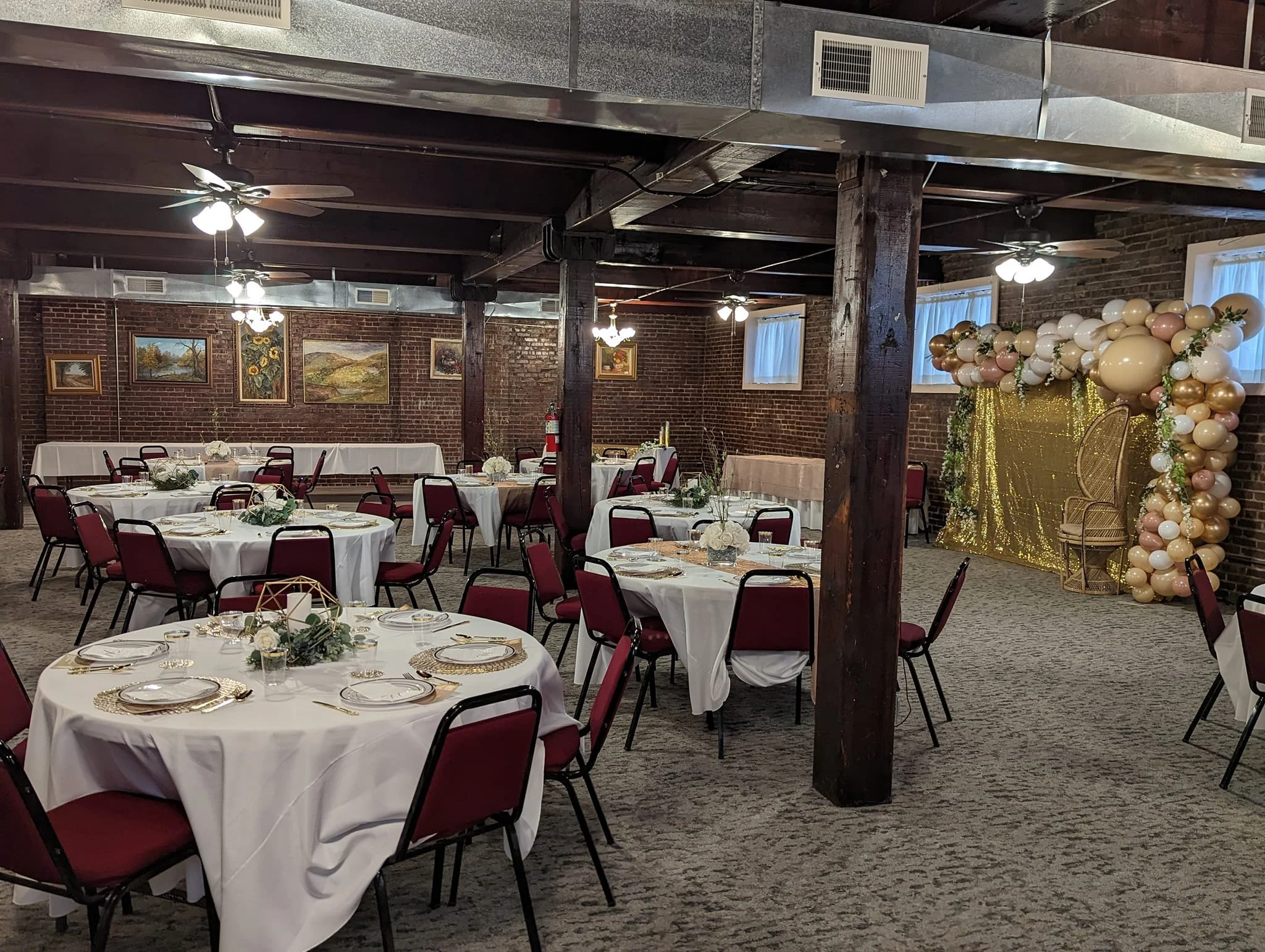 Event space decorated with round tables draped in white tablecloths, red chairs, floral centerpieces, and a balloon arch with gold, white, and blush balloons and gold sequin backdrop.