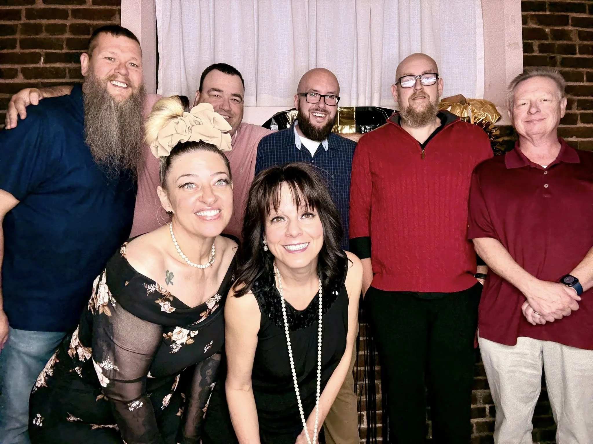 Group of eight people posing indoors in front of brick wall and curtains, smiling at the camera.