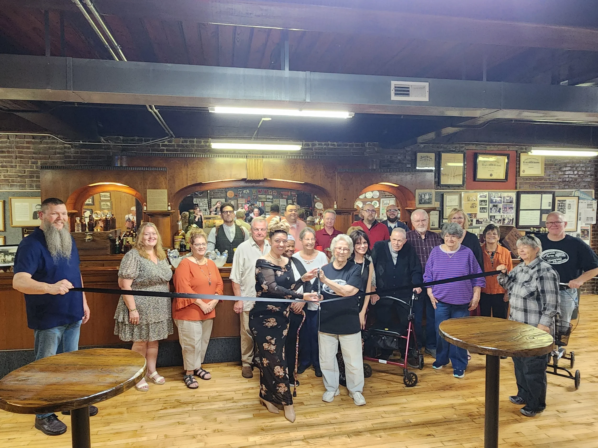 A group of people gathered in a restaurant or pub, some holding a ribbon for a ribbon-cutting ceremony, with a bar in the background and framed photos and awards on the wall.