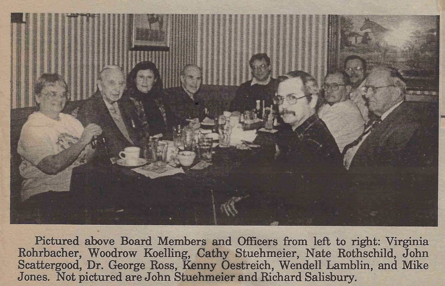 A black-and-white photo of eleven people sitting around a long table in a restaurant or dining room, with food and drinks on the table. The people are smiling and looking at the camera. There are framed pictures on the wall behind them.