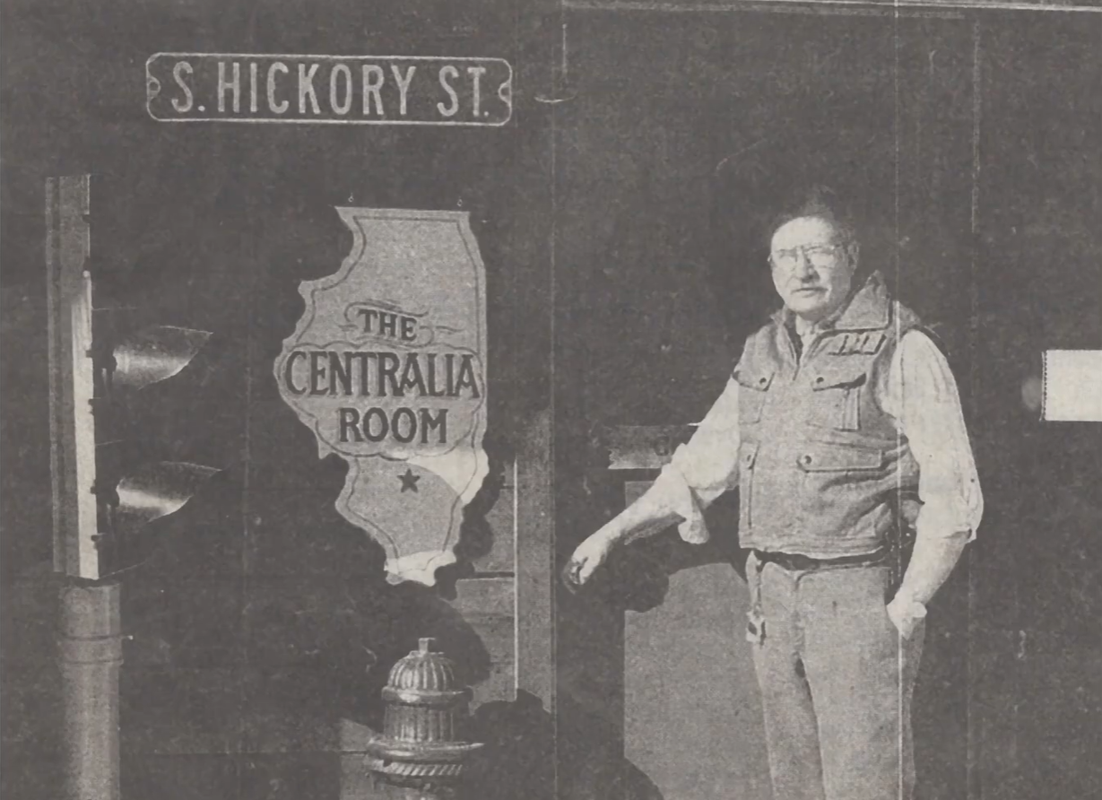 A man wearing glasses and a vest standing next to a sign that says "The Centralia Room" and a map of Illinois with a star indicating a location, with a street sign that reads "S. Hickory St."