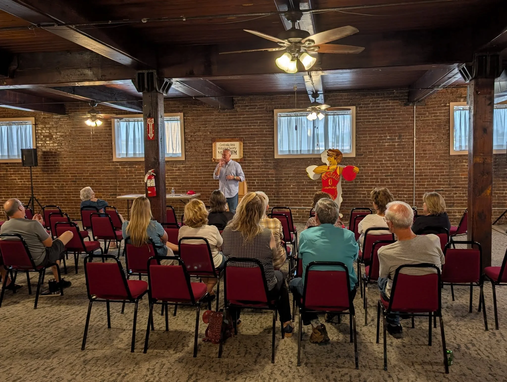 A man giving a presentation to a small audience in a brick-walled room with red chairs, windows, ceiling fans, and a New Mexico-themed decorations.