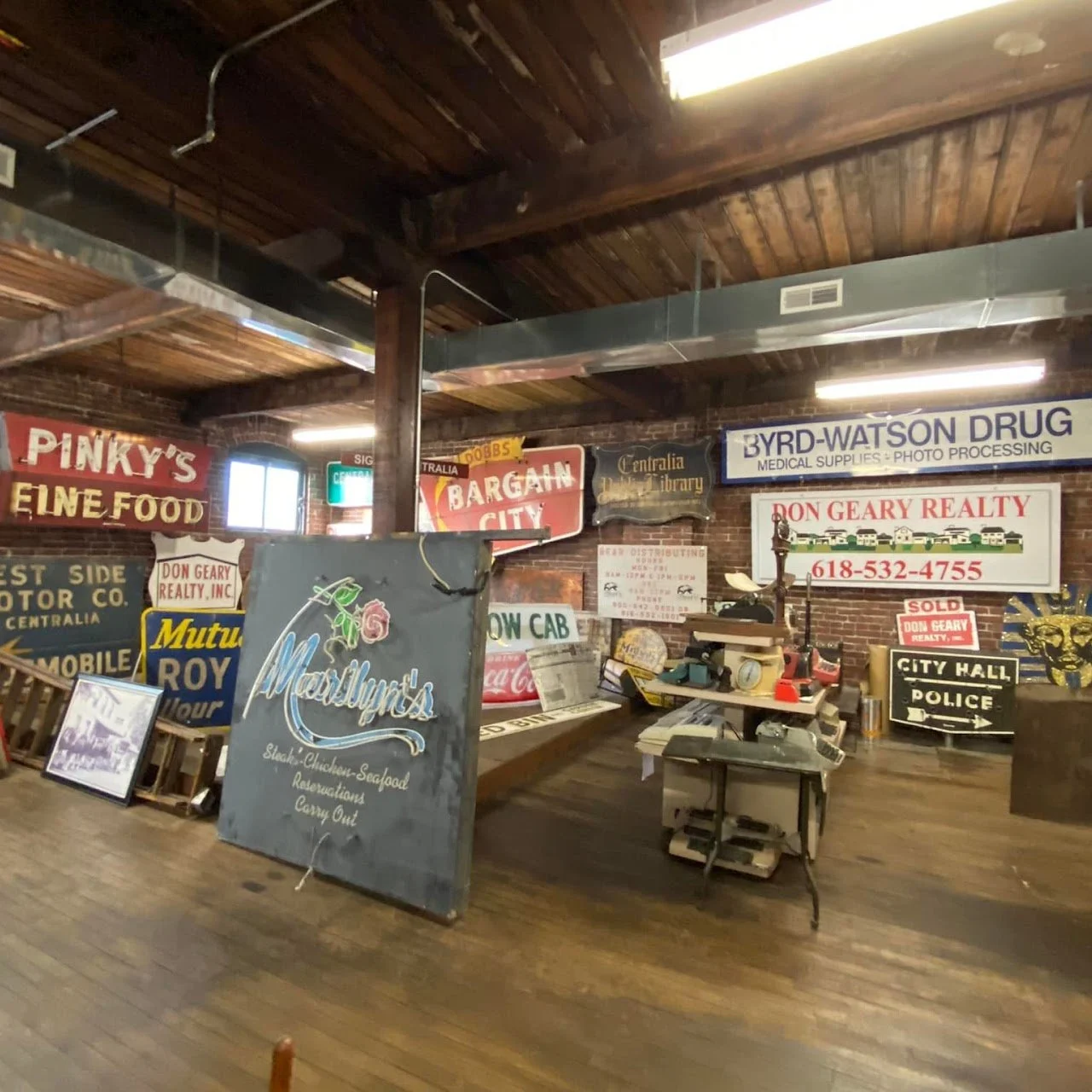 Interior of an antique shop or auction house filled with vintage signs, posters, and memorabilia, including business signs for drugstores, realty, city hall, police, and various businesses.