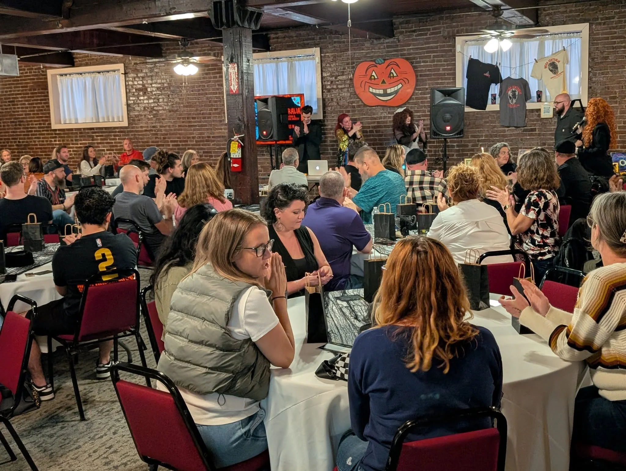 Indoor event with a large group of people seated at tables, watching a performance or presentation on a stage. The decor includes Halloween-themed decorations, such as a large pumpkin face on the wall, and wrapped gift bags on the tables. Some audience members are clapping or observing attentively.