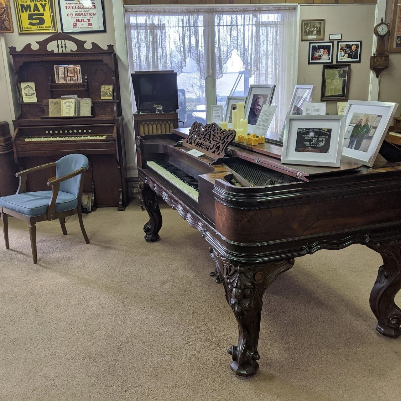 Room with a vintage grand piano decorated with framed photographs and candles, a wood cabinet with books and a small piano, and a chair with a blue cushion, all set against lace curtains and framed wall art.